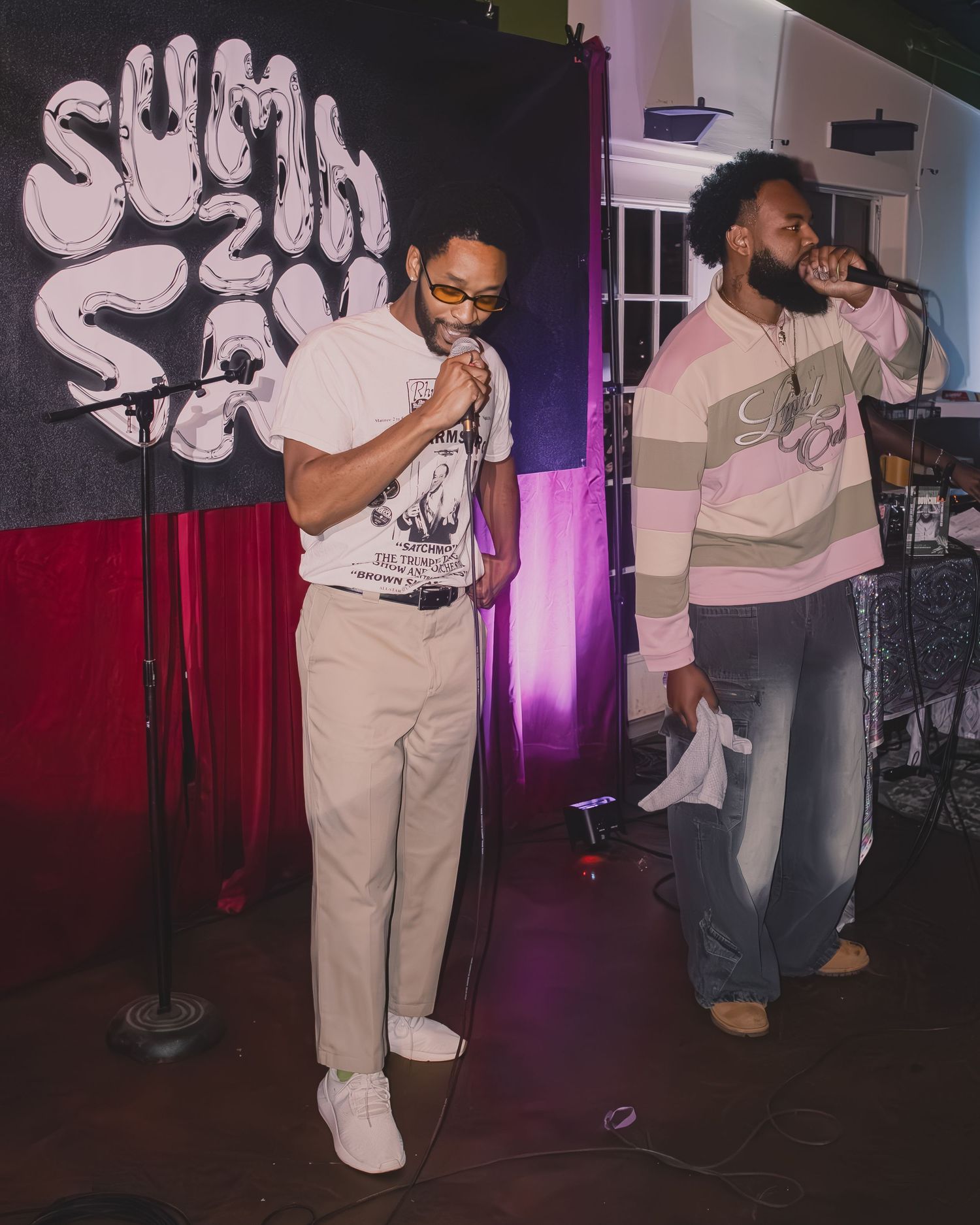 Two performers on stage with Sumi and Say banner backdrop in matching light-colored outfits.