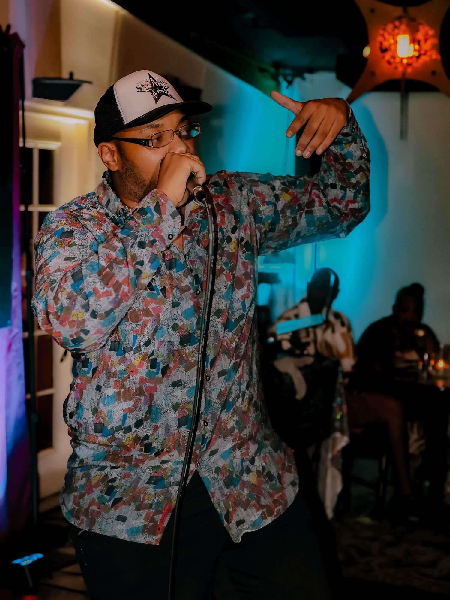 A performer in a patterned shirt and baseball cap gestures energetically during a live performance with blue lighting.
