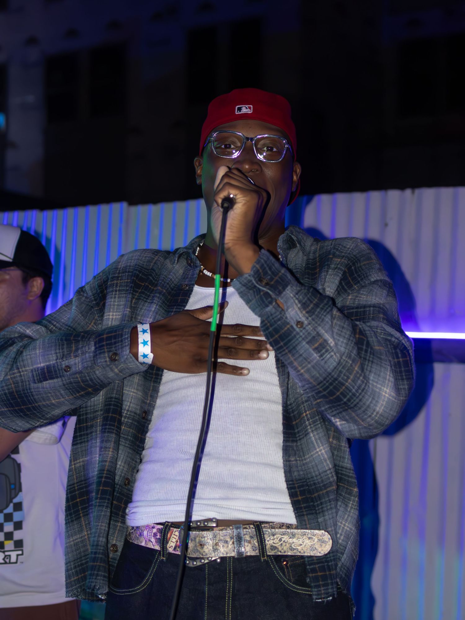 Artist performing on stage wearing plaid jacket and glasses under blue stage lights.