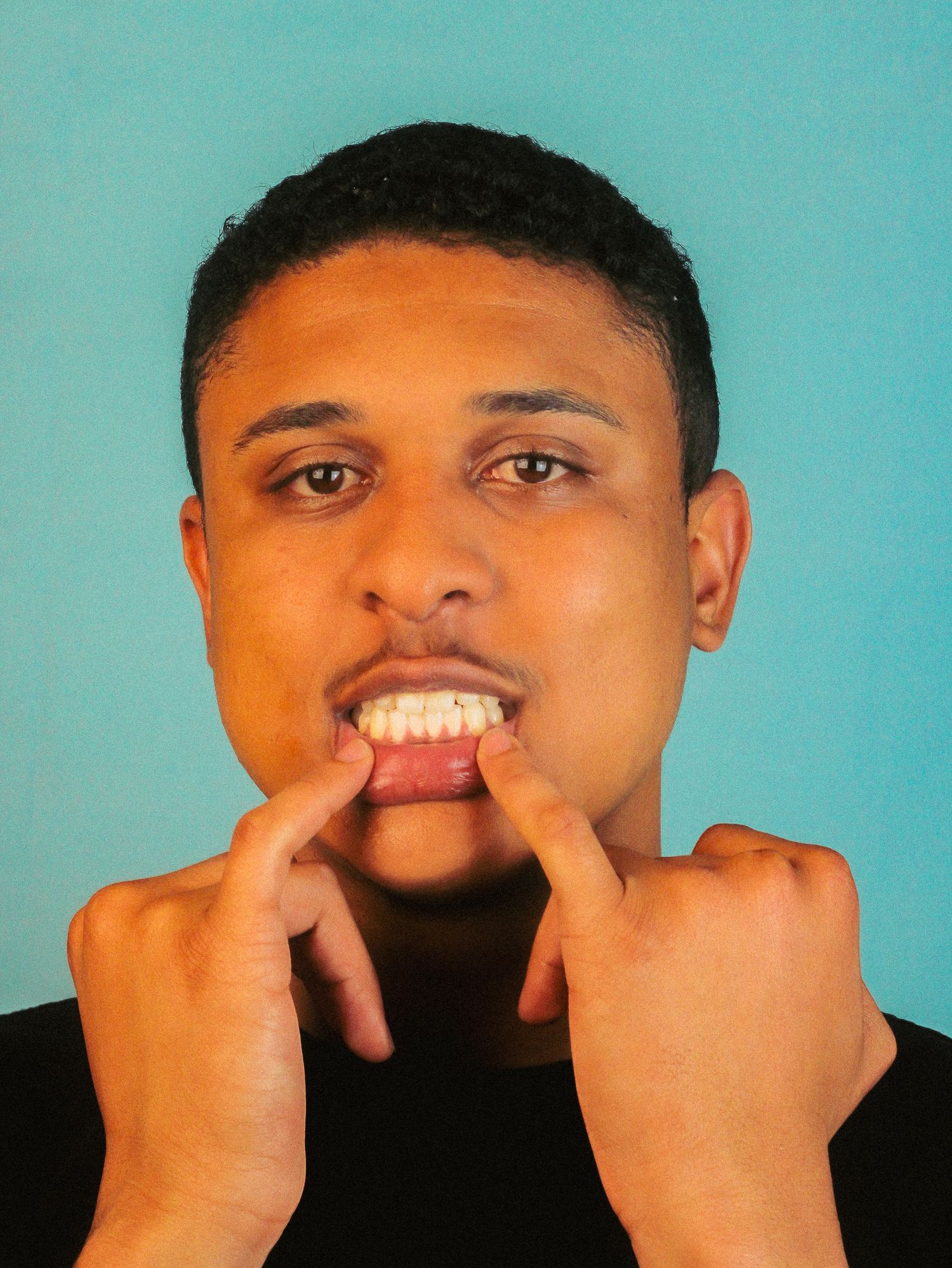 Young man in black shirt pulling corners of mouth wide open against turquoise background.
