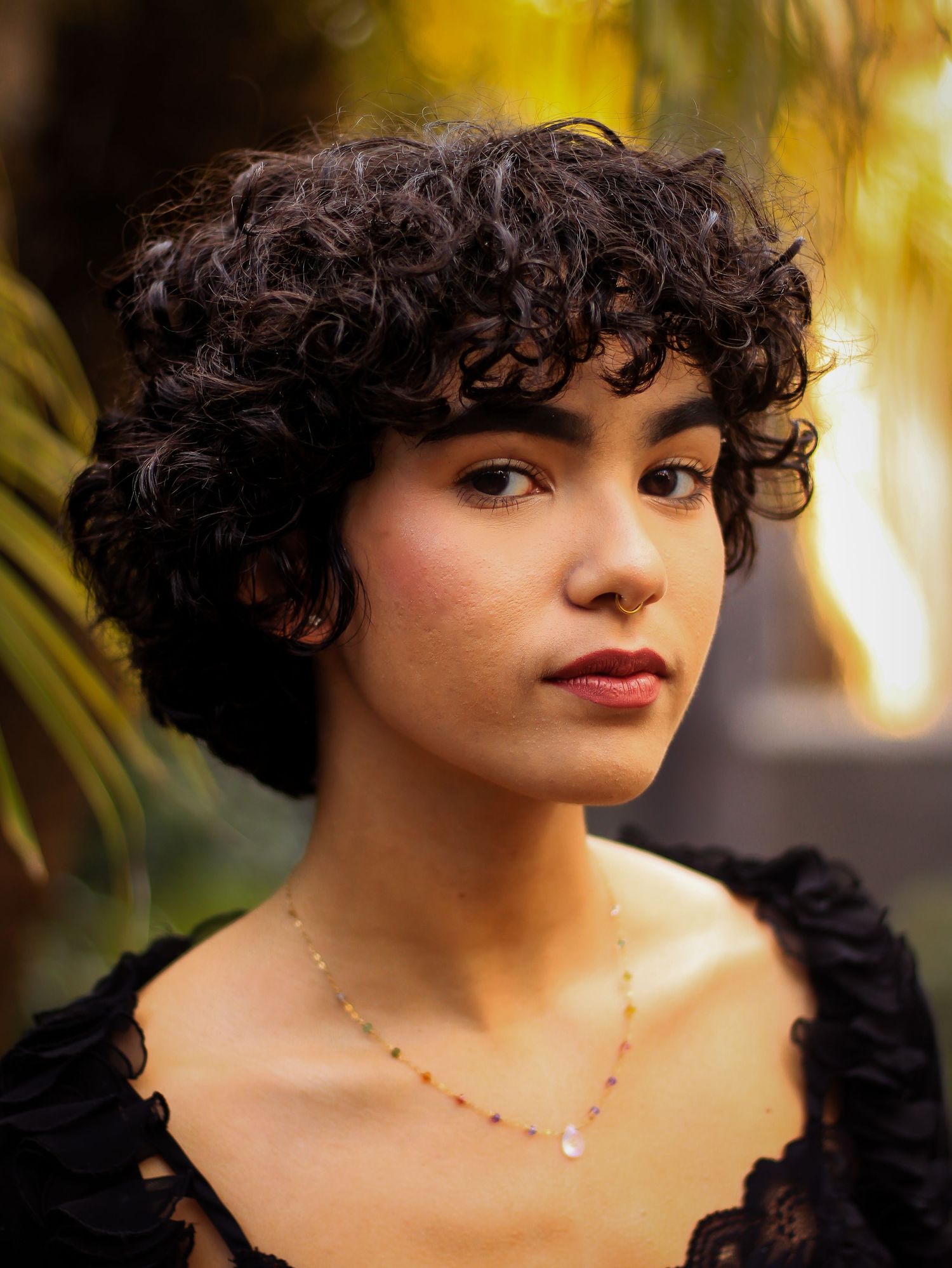 Artistic outdoor portrait with curls and delicate necklace against warm lighting.