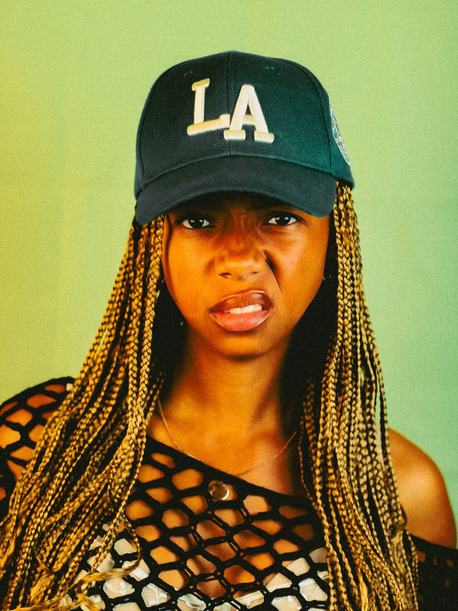 Young woman wearing LA baseball cap and mesh top poses with neutral expression against green background.