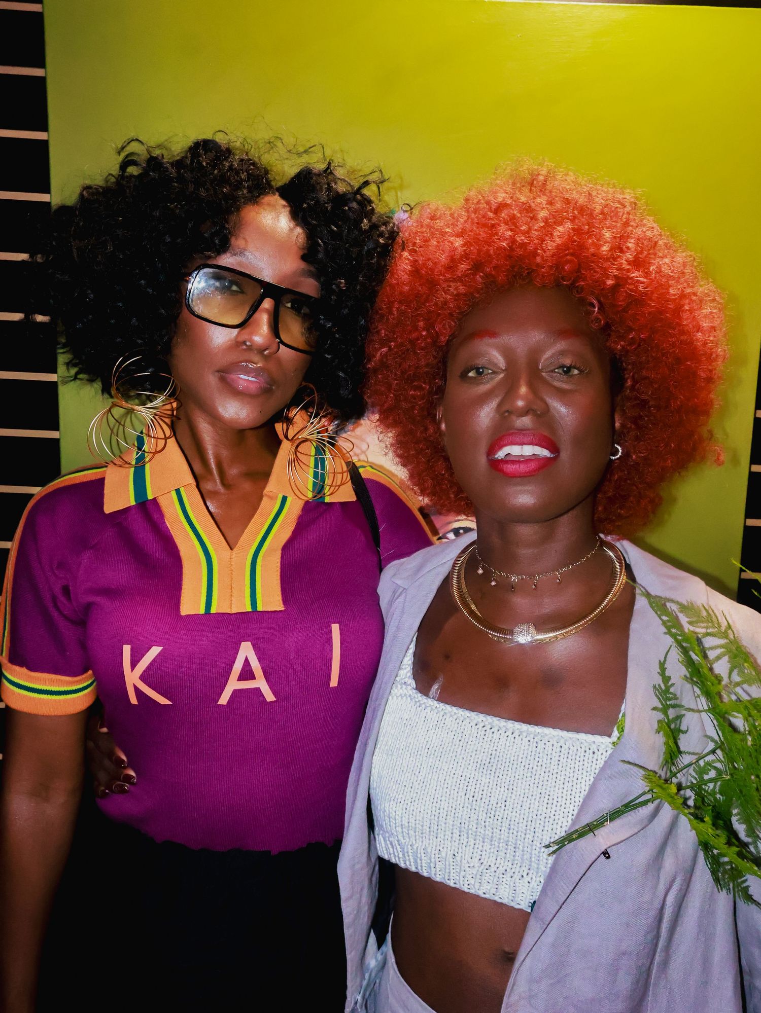 Two friends pose together against a yellow wall wearing purple and white outfits.