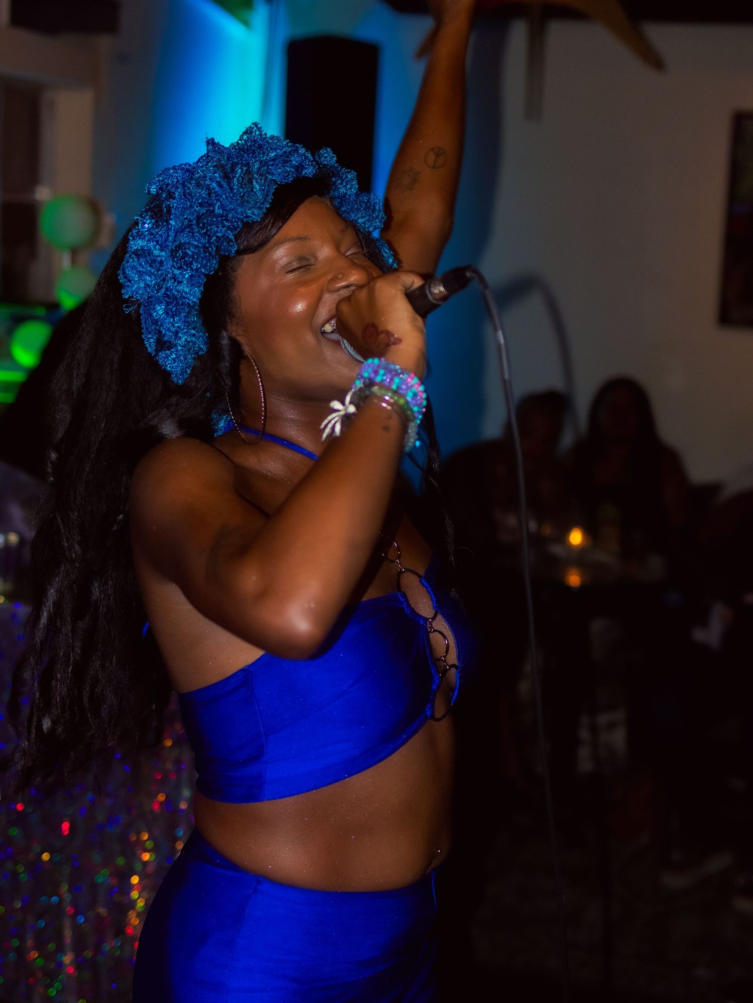 Performer in a blue outfit and matching blue wig sings passionately under dramatic lighting at a nightclub venue.