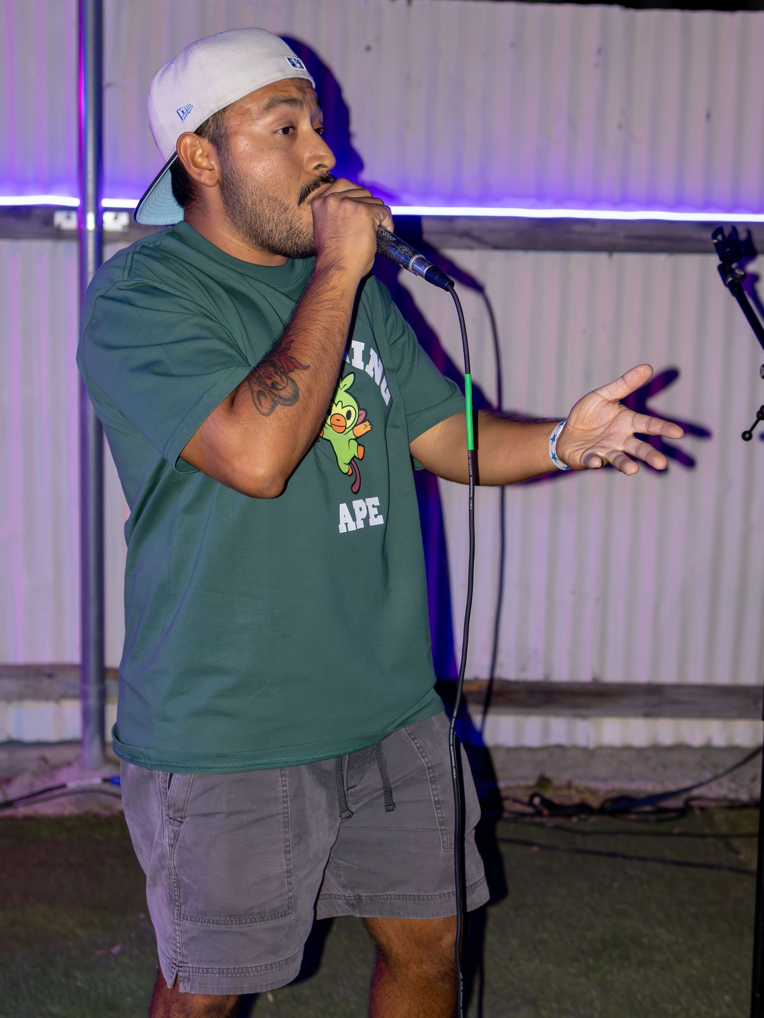 Performer in green shirt and white cap gesturing while speaking into microphone on stage.