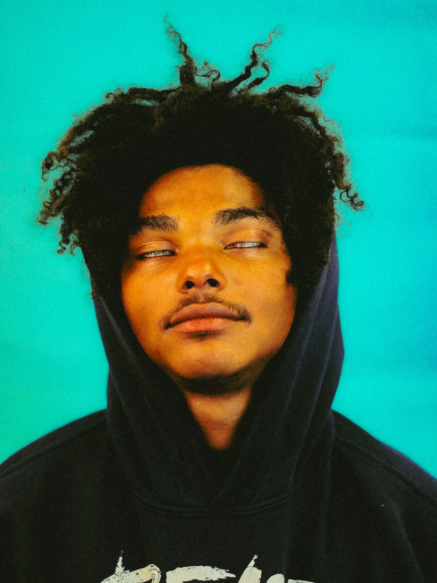Young man with curly hair wearing black hoodie with eyes closed against turquoise background.