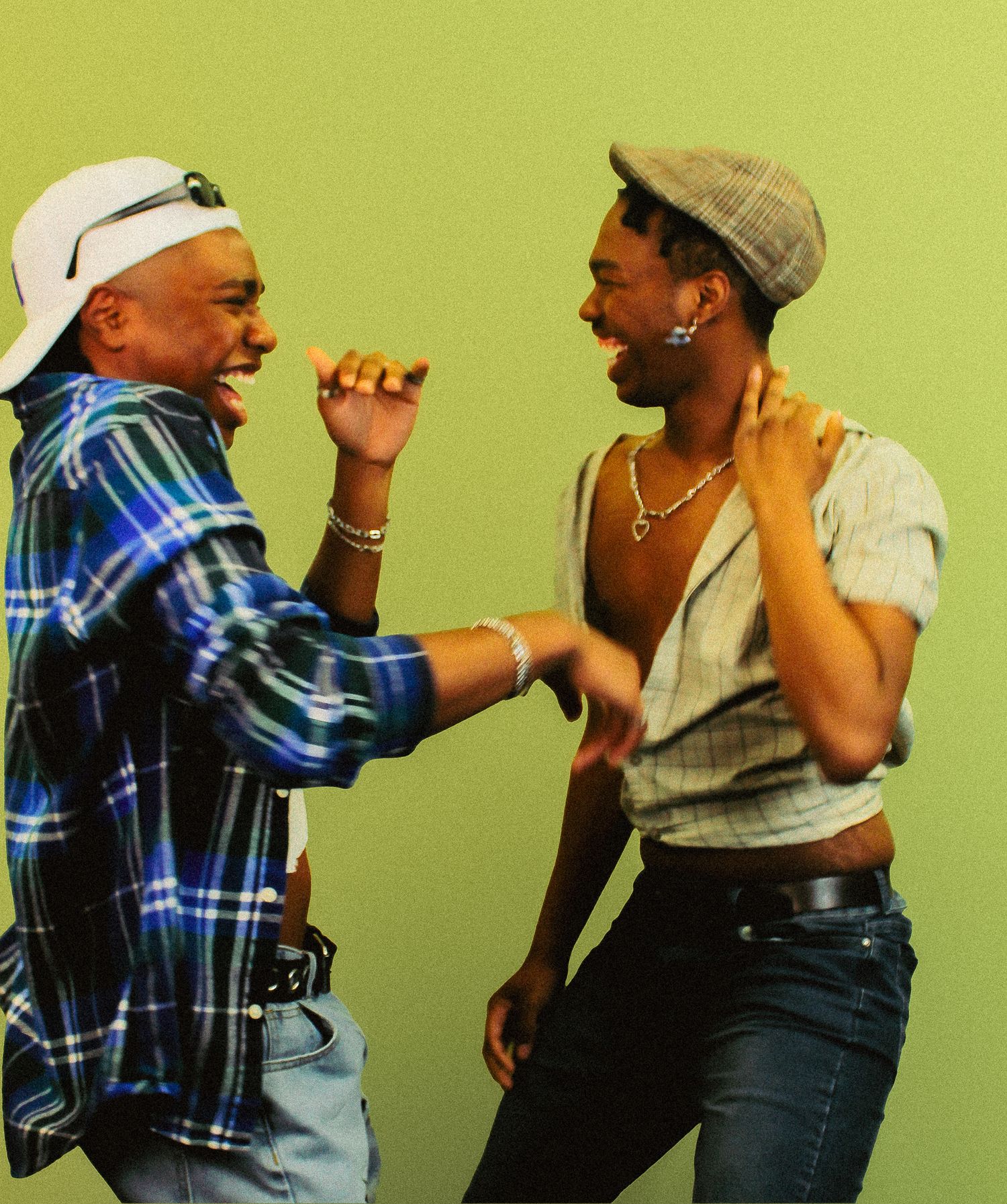 Two young men laugh and dance together wearing flannel and plaid shirts against a green background.