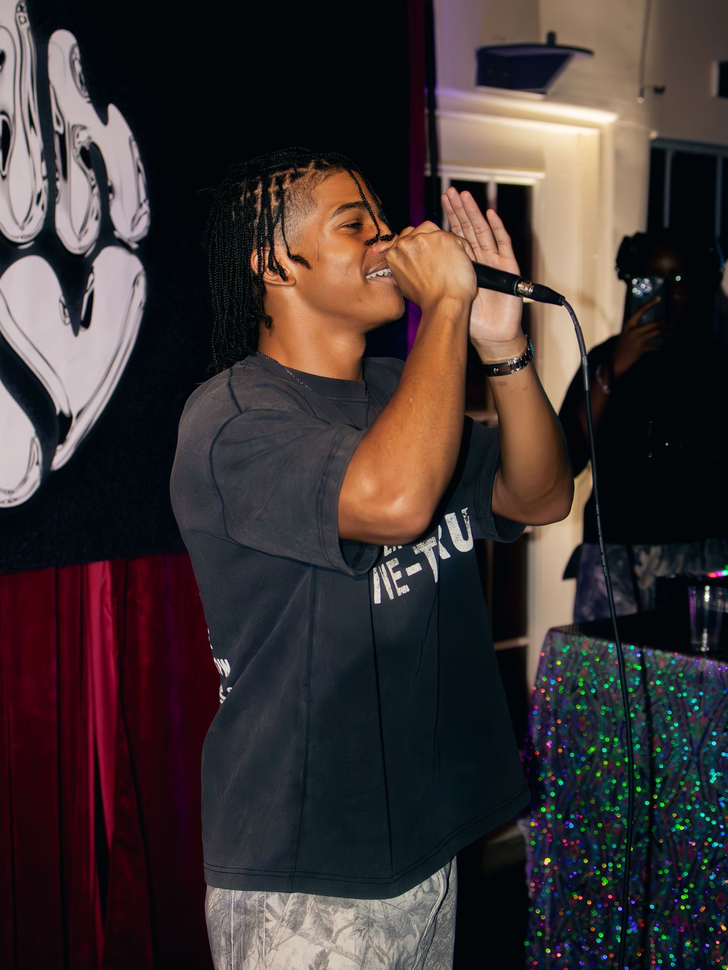 Singer performing on stage with a microphone against a backdrop of colorful lights and wall decorations.