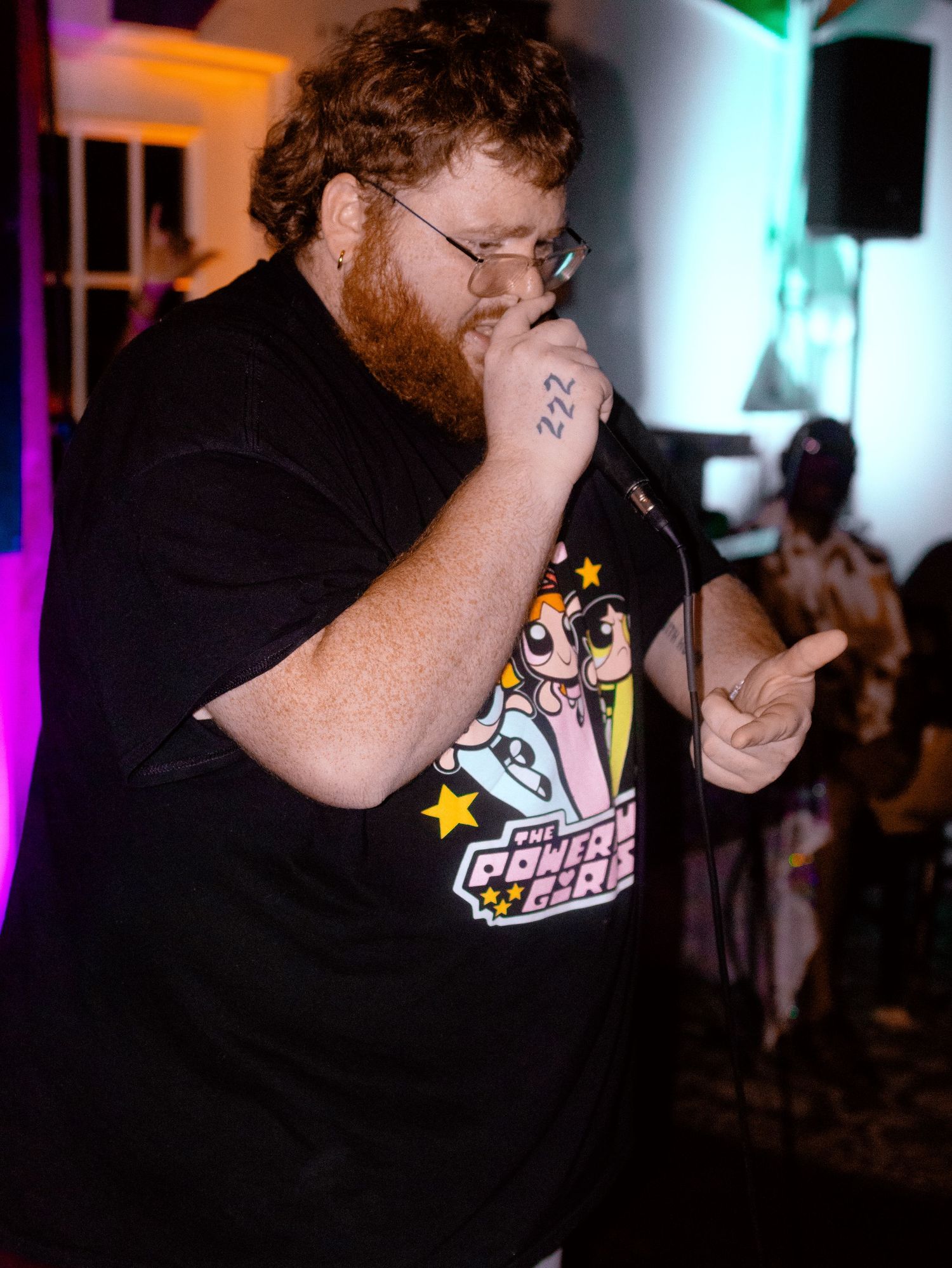 Performer in a black t-shirt with colorful graphics singing into a microphone at a dimly lit venue.