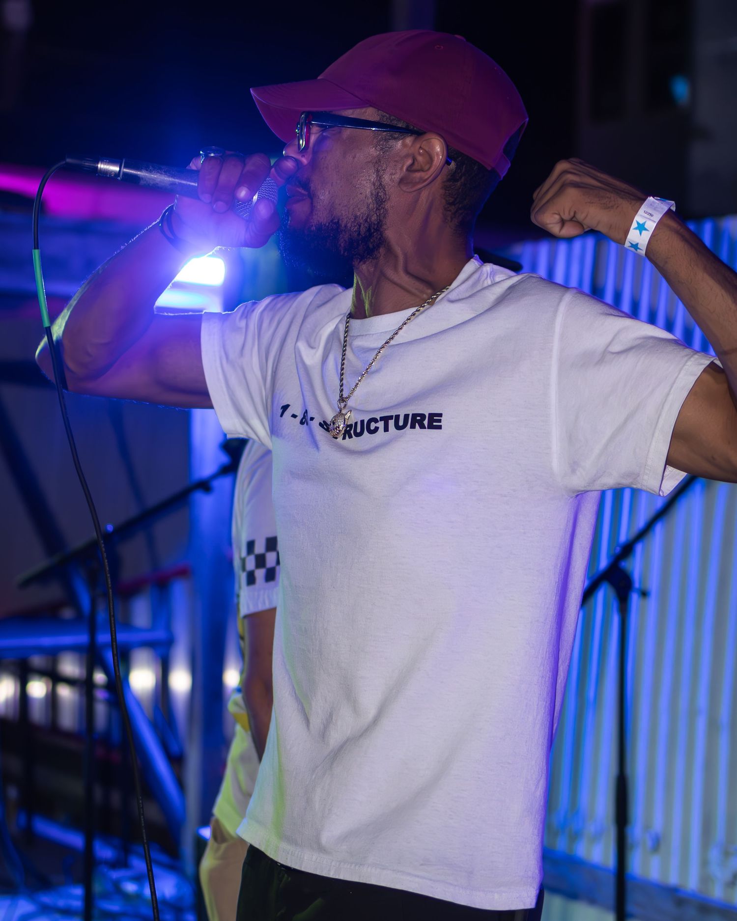 Performer in white t-shirt and purple cap singing on stage with blue and purple lighting effects.