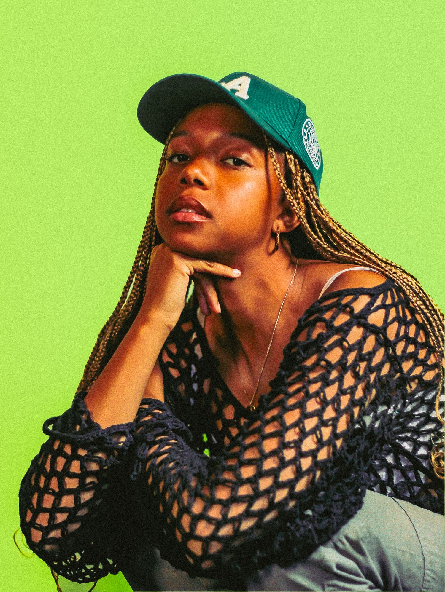 Young woman in green cap and black mesh top posing against a vivid green background.