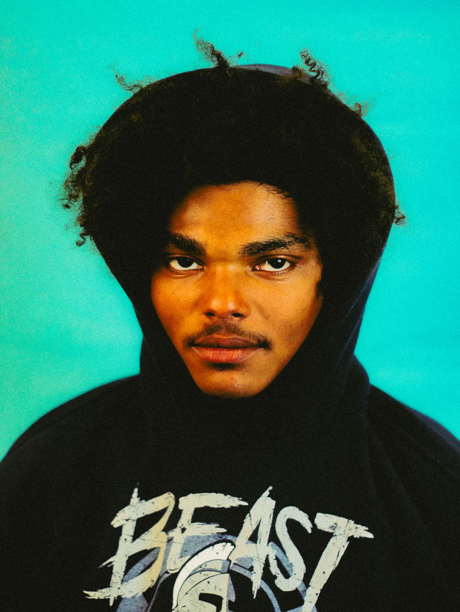 Young man wearing black Beast hoodie with curly hair poses against vibrant teal background.