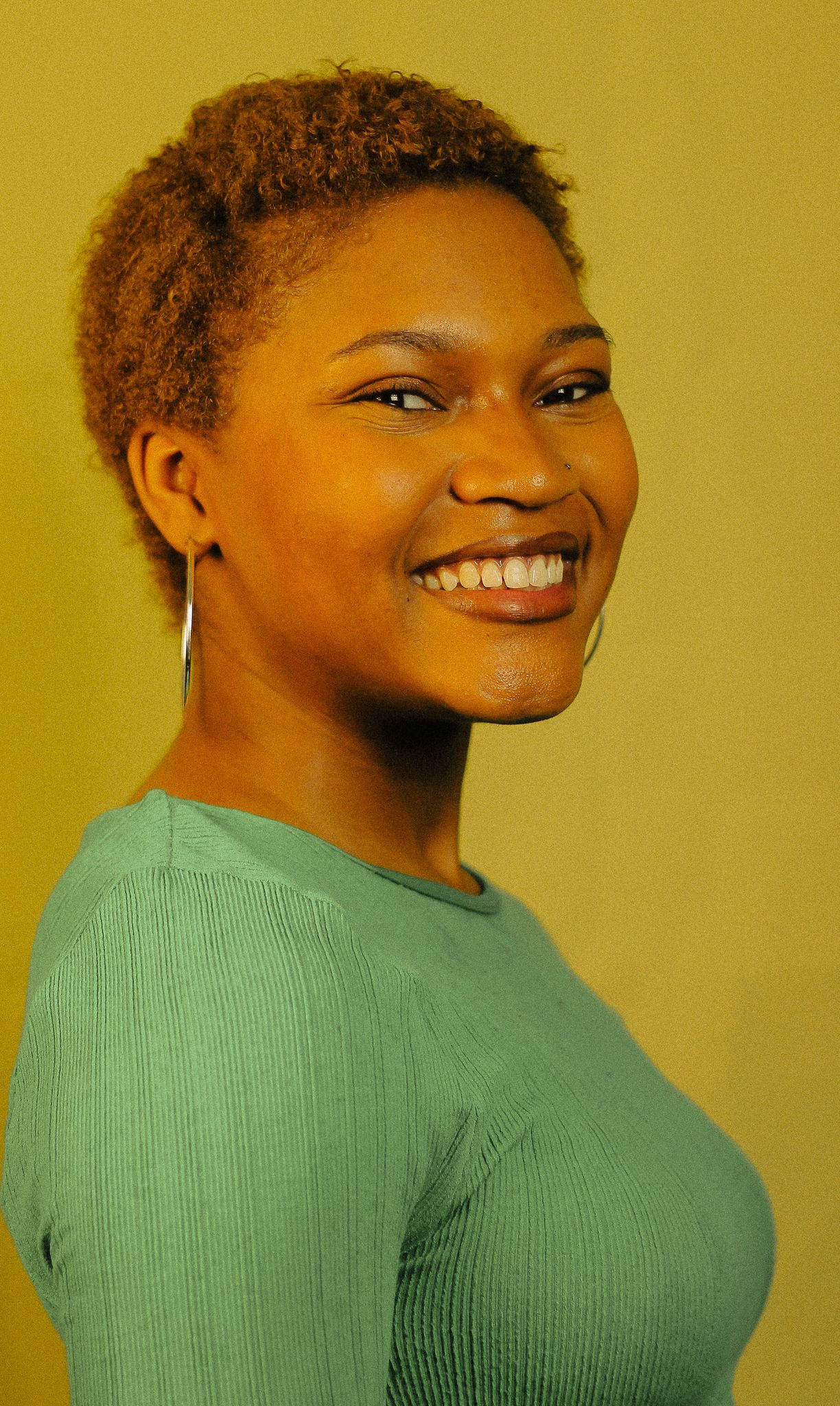 Young woman with short golden natural hair smiling sideways, wearing teal ribbed top and hoop earring.