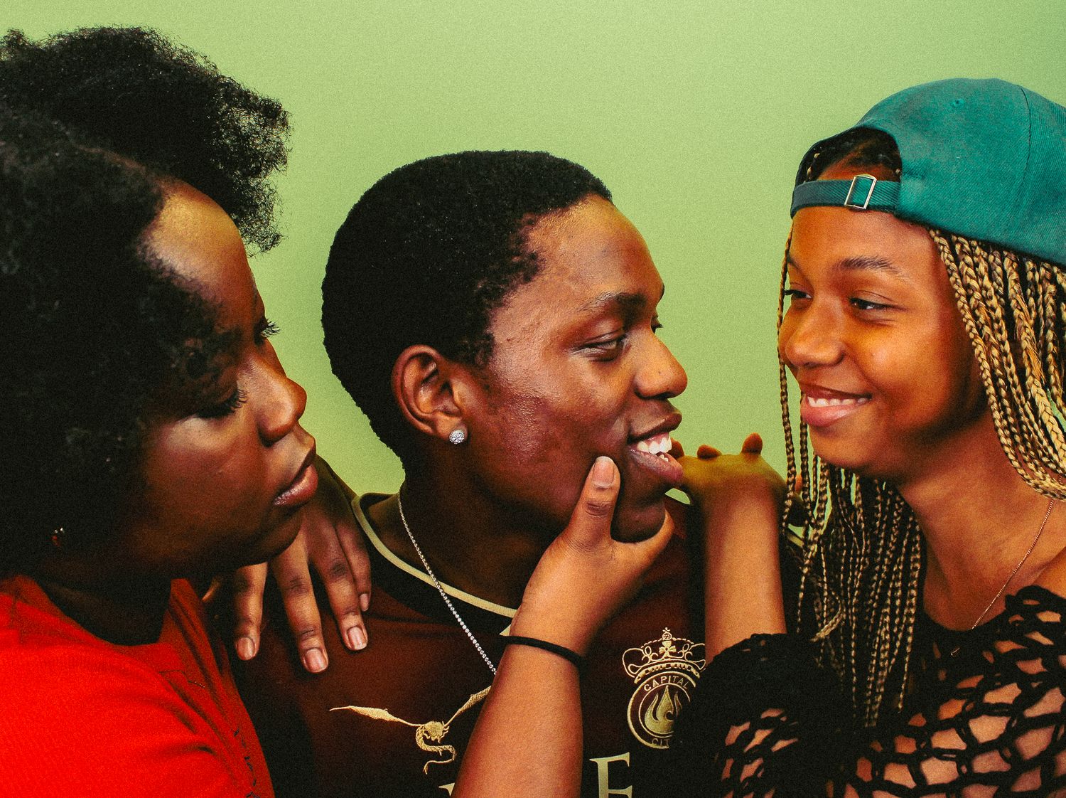 Three young Black friends share a joyful moment together, laughing and touching faces against a green background.