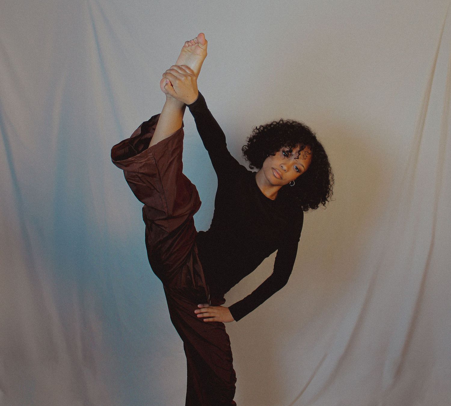 A dancer performs fluid movements in brown clothing against a white backdrop.