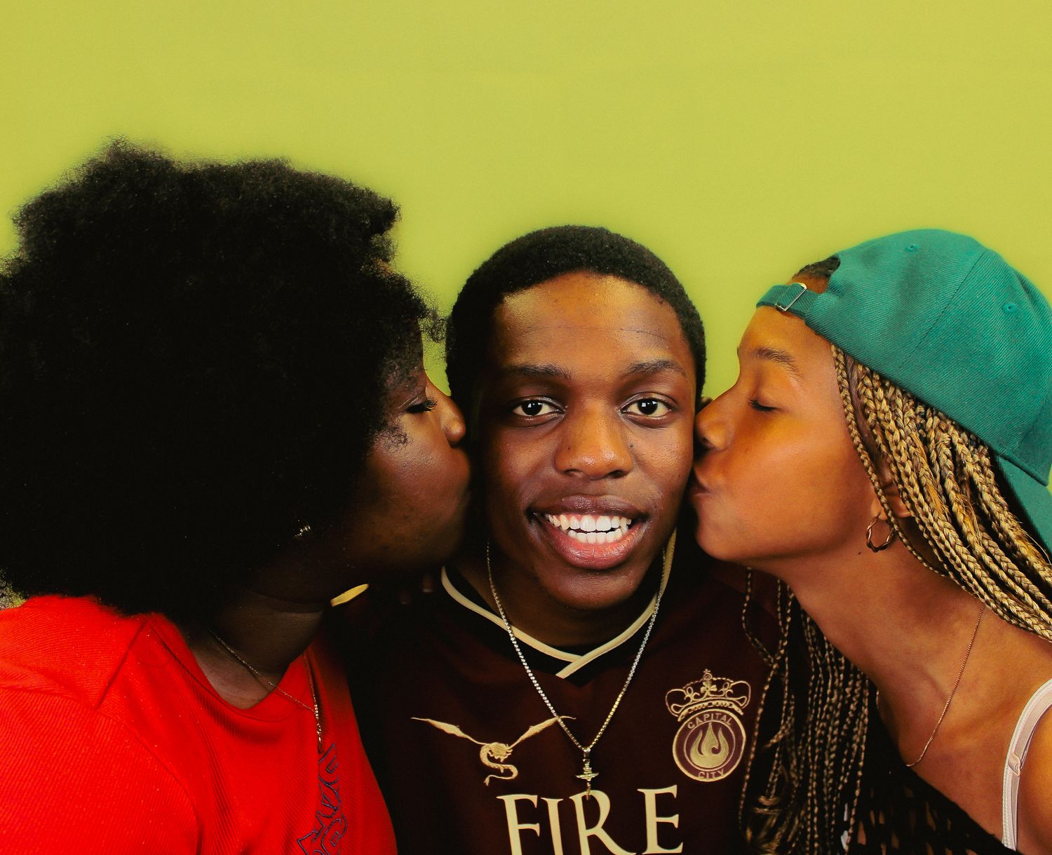 Three young people pose together against a green wall, with two women kissing a smiling young man on each cheek.