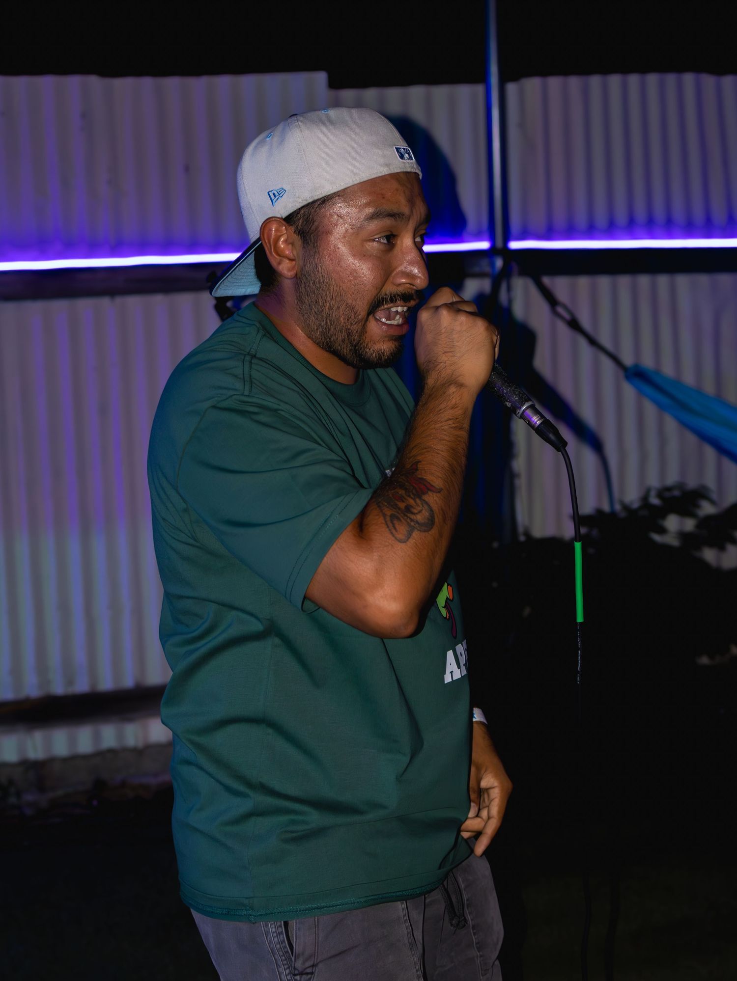 Performer in green shirt and white cap speaks into microphone with purple lighting in background.
