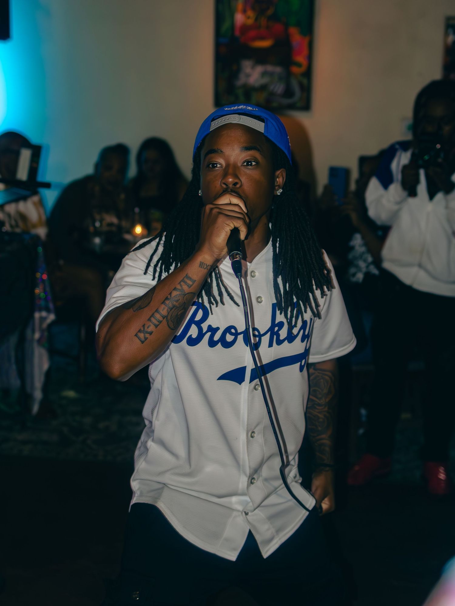 Artist in Brooklyn baseball jersey performing at venue with blue ambient lighting.
