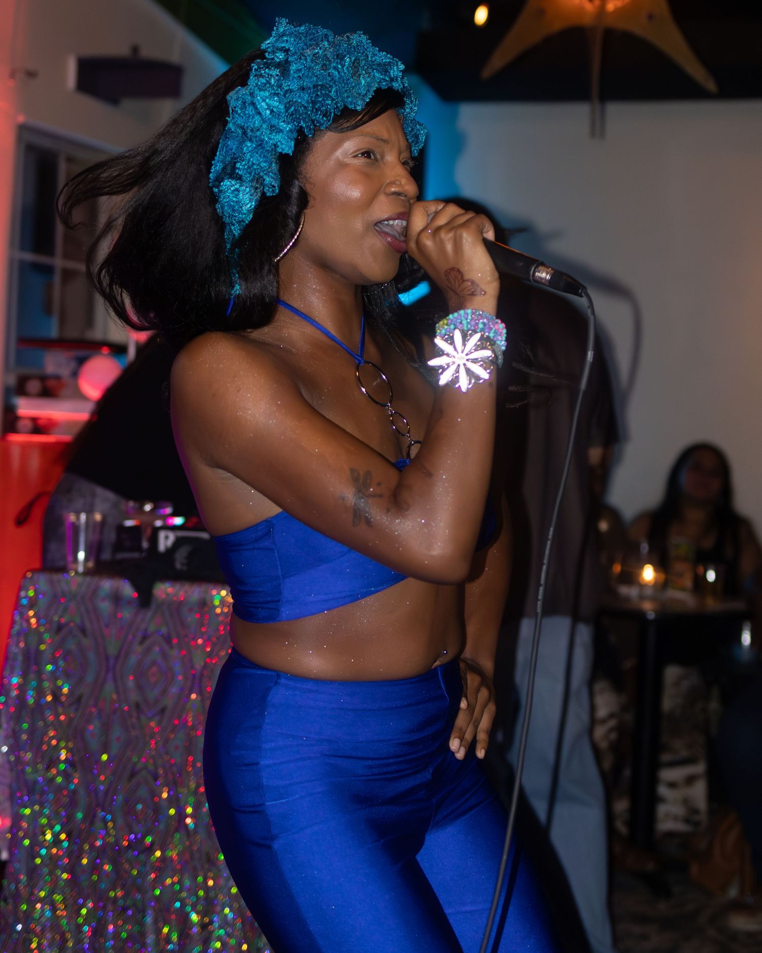 Performer in blue outfit and wig sings passionately under multicolored stage lights.
