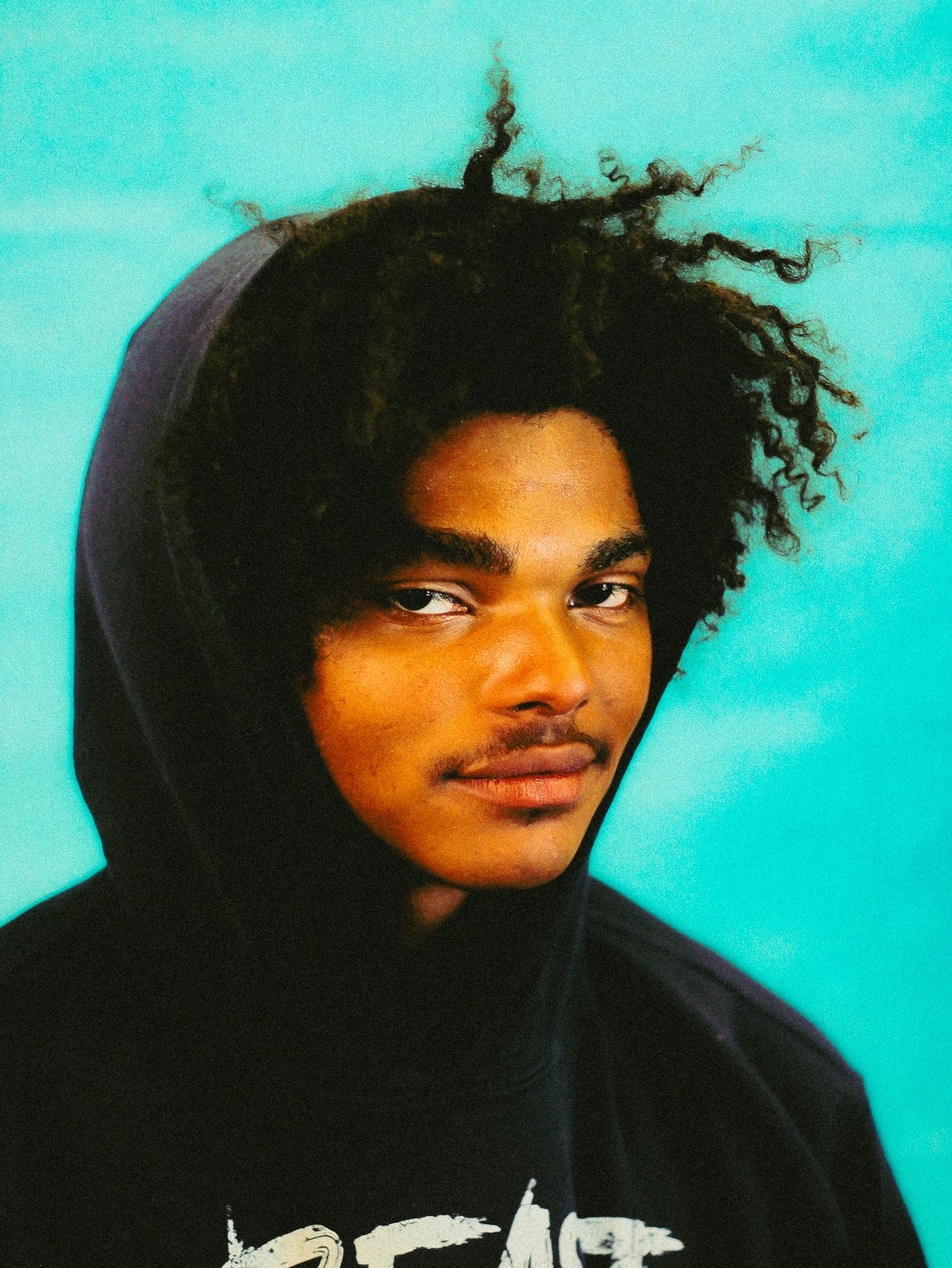 Young man with curly hair wearing black hoodie posing seriously against turquoise background.