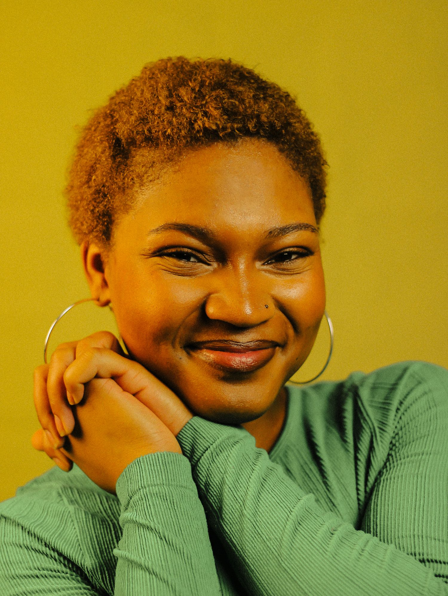 A young Black woman with short natural hair smiles warmly wearing hoop earrings against a yellow background.