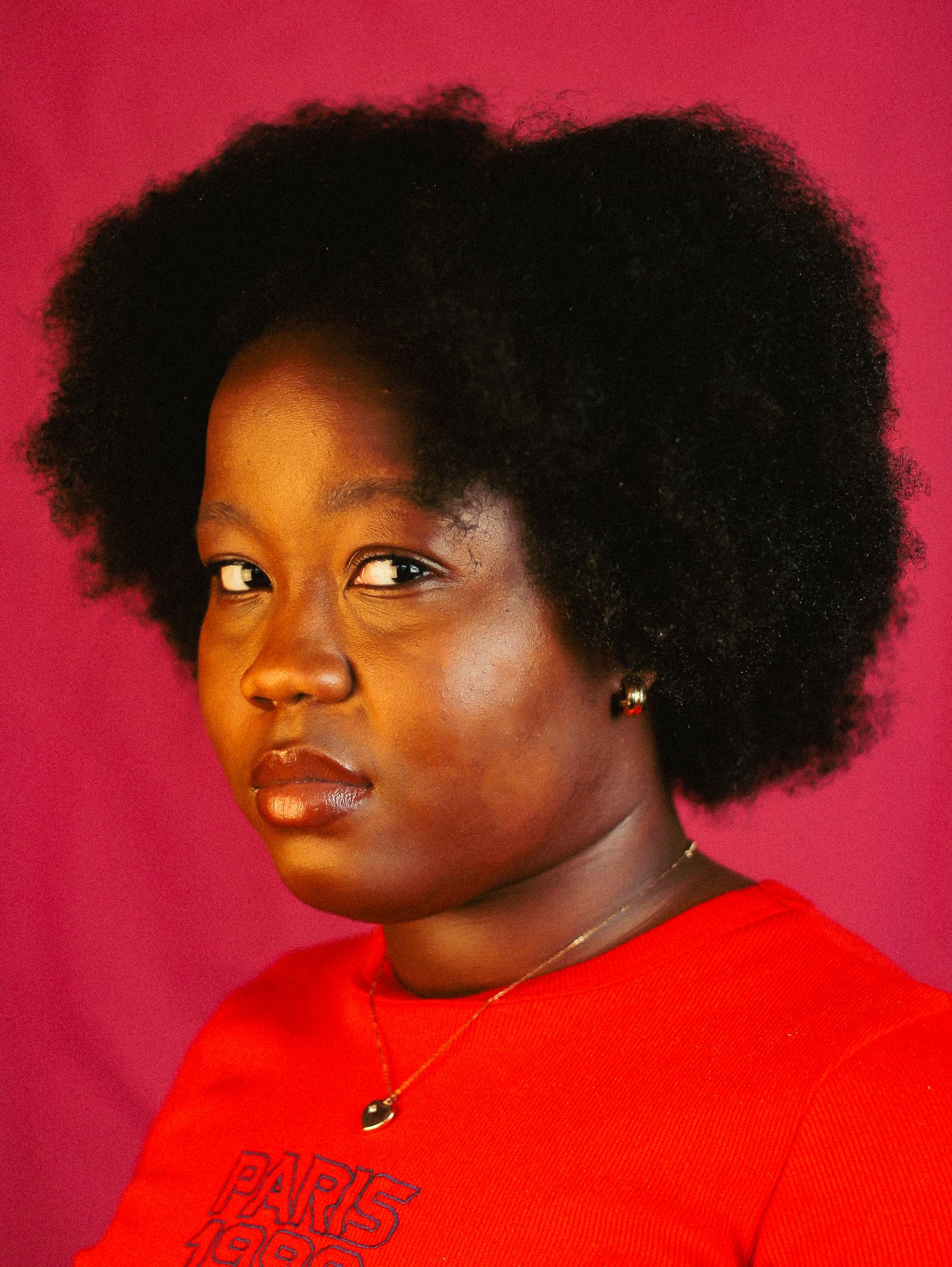 Black woman with large natural afro hair wearing a red Paris 1999 shirt, looking sideways on pink background.