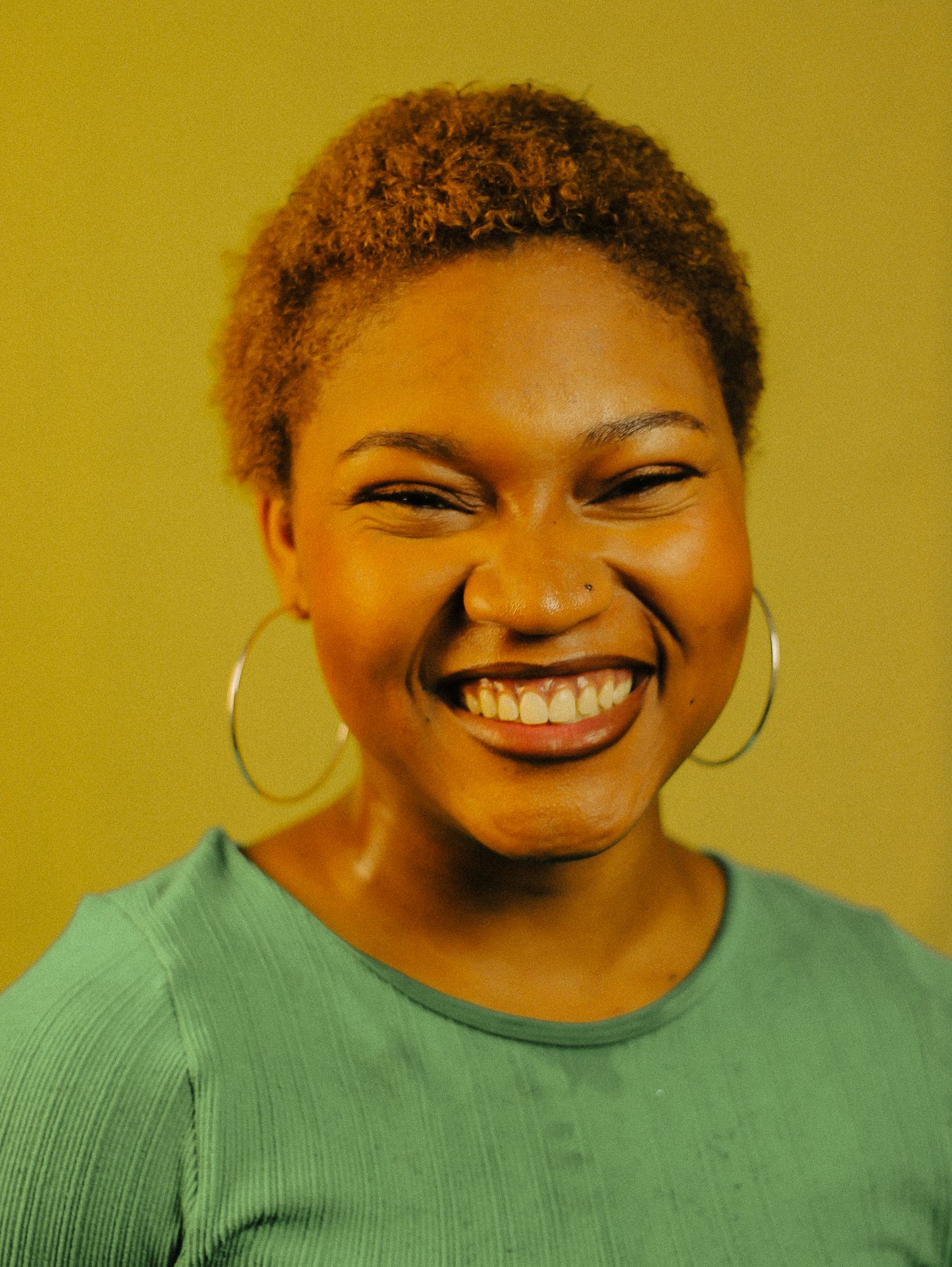 A young Black woman with short natural hair laughs joyfully wearing large hoop earrings on a yellow background.