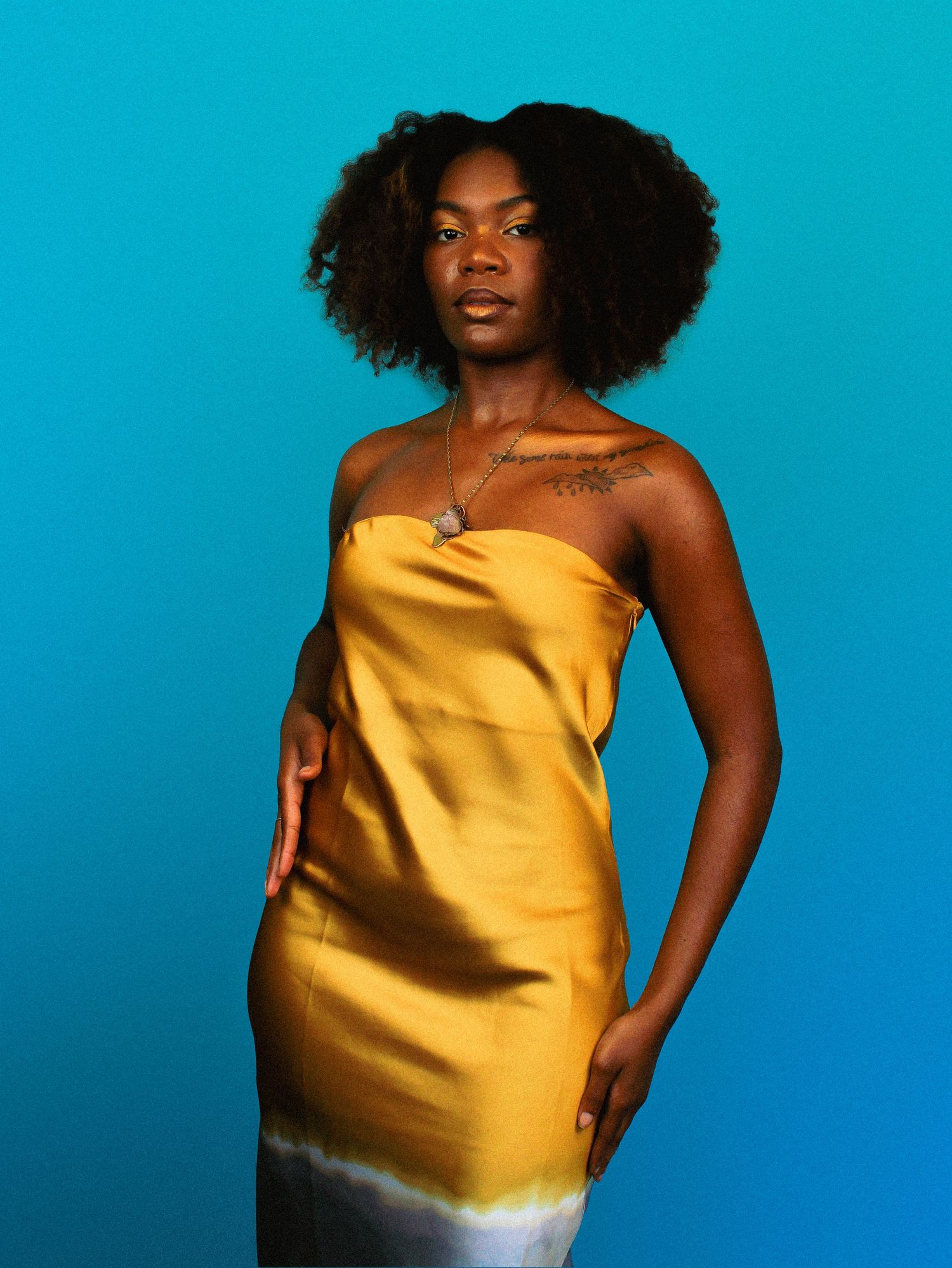 Model in gold satin dress poses confidently against bright turquoise background.