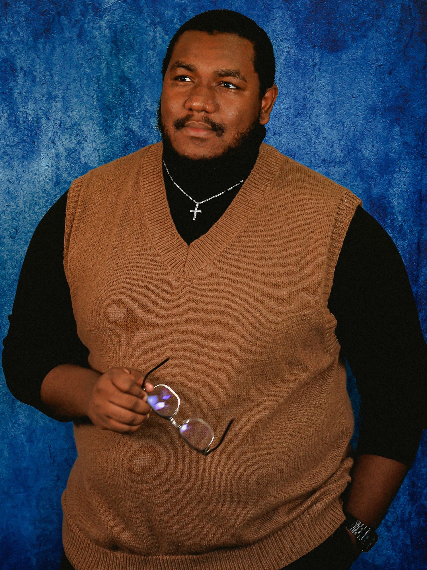 Person in brown sweater vest and black shirt poses against blue backdrop.