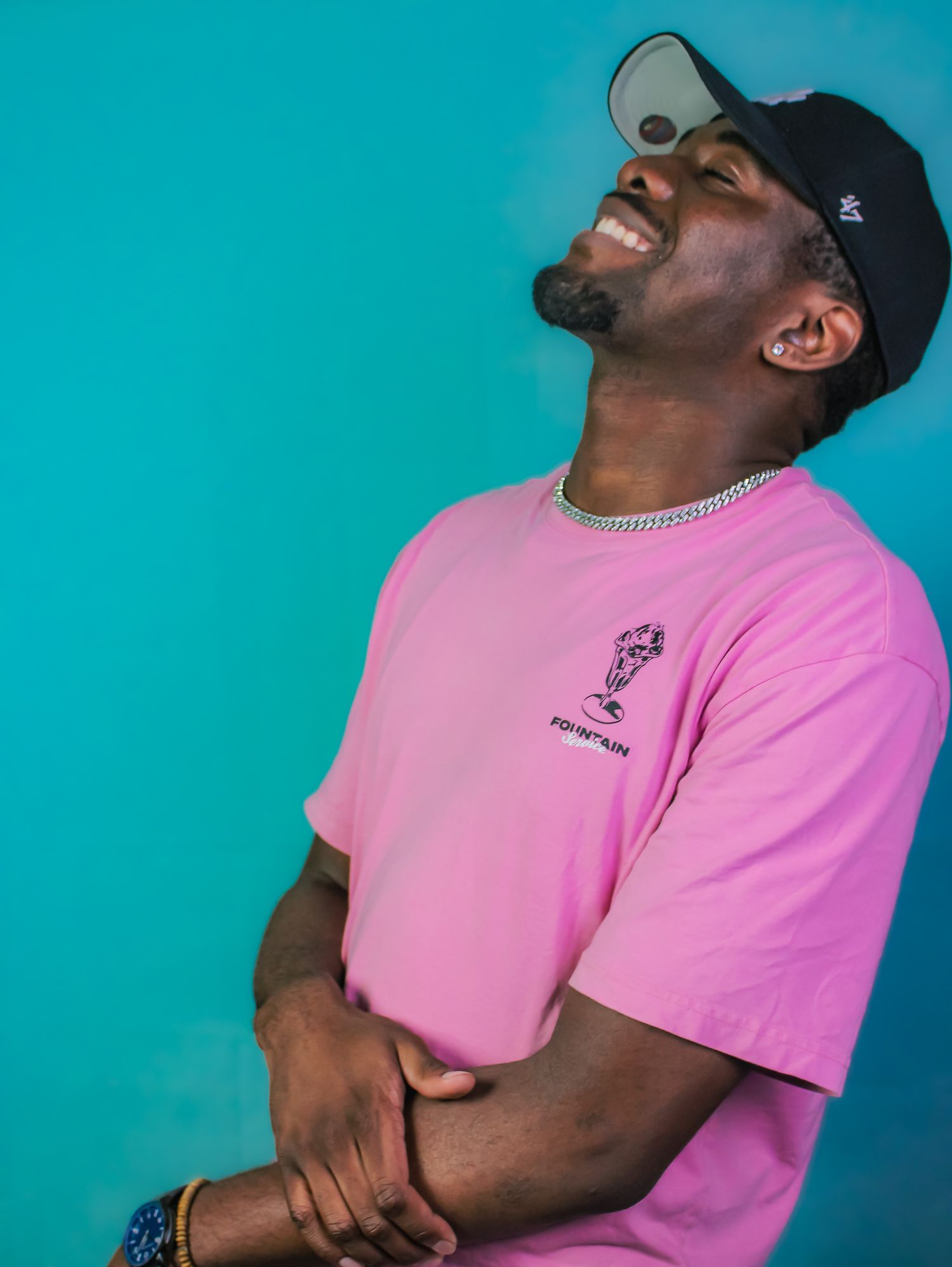 A smiling man in a pink t-shirt and black cap laughs joyfully against a teal background.