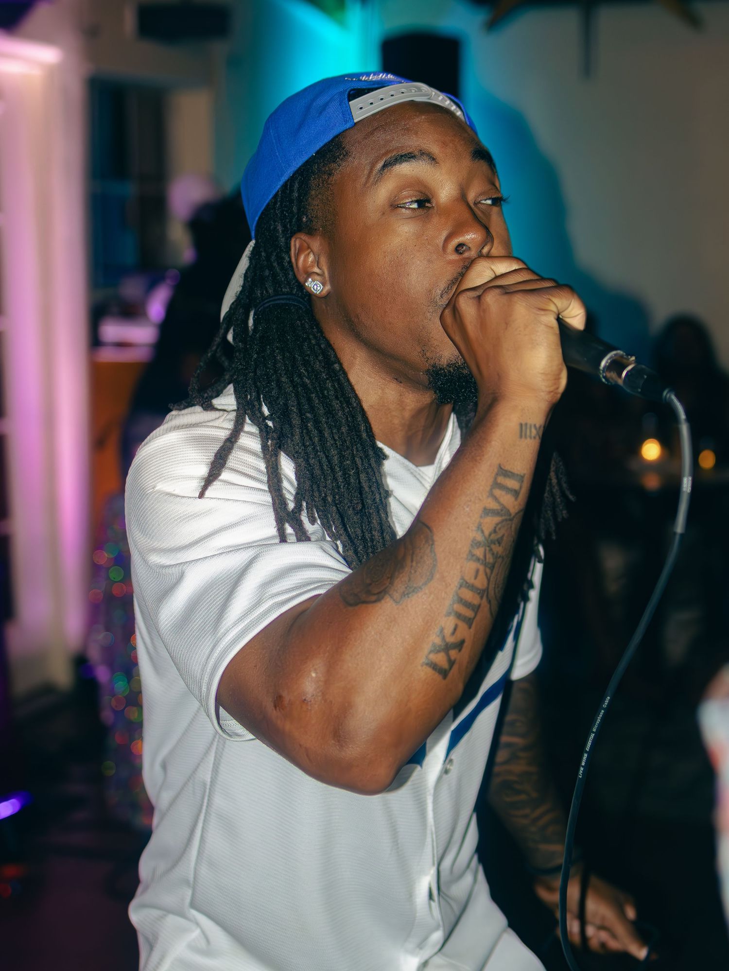 Rapper with dreadlocks performs passionately in white jersey and blue cap under colorful lights.