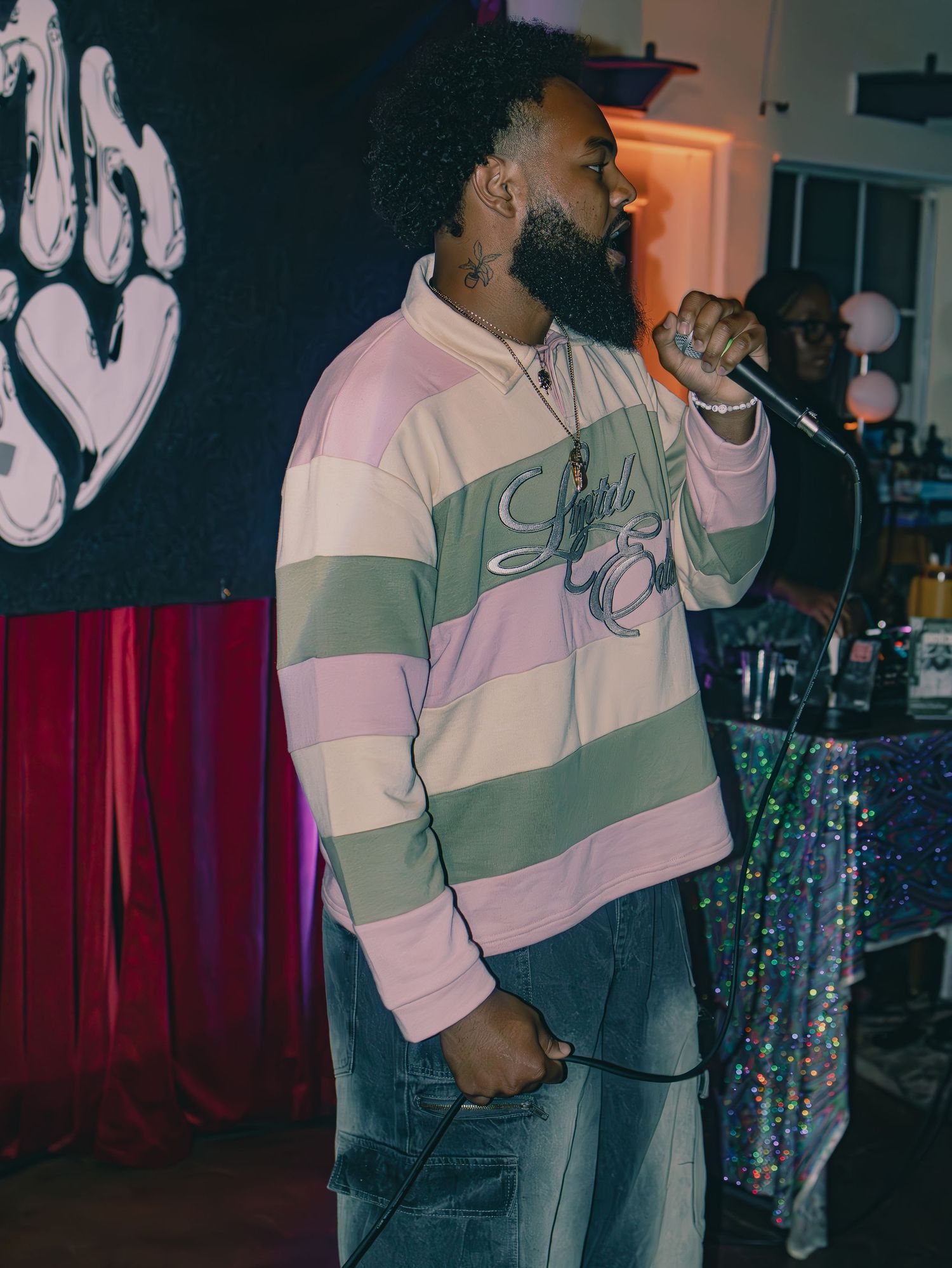 A performer in a white and gray sweatshirt singing into a microphone on stage with colorful lighting.
