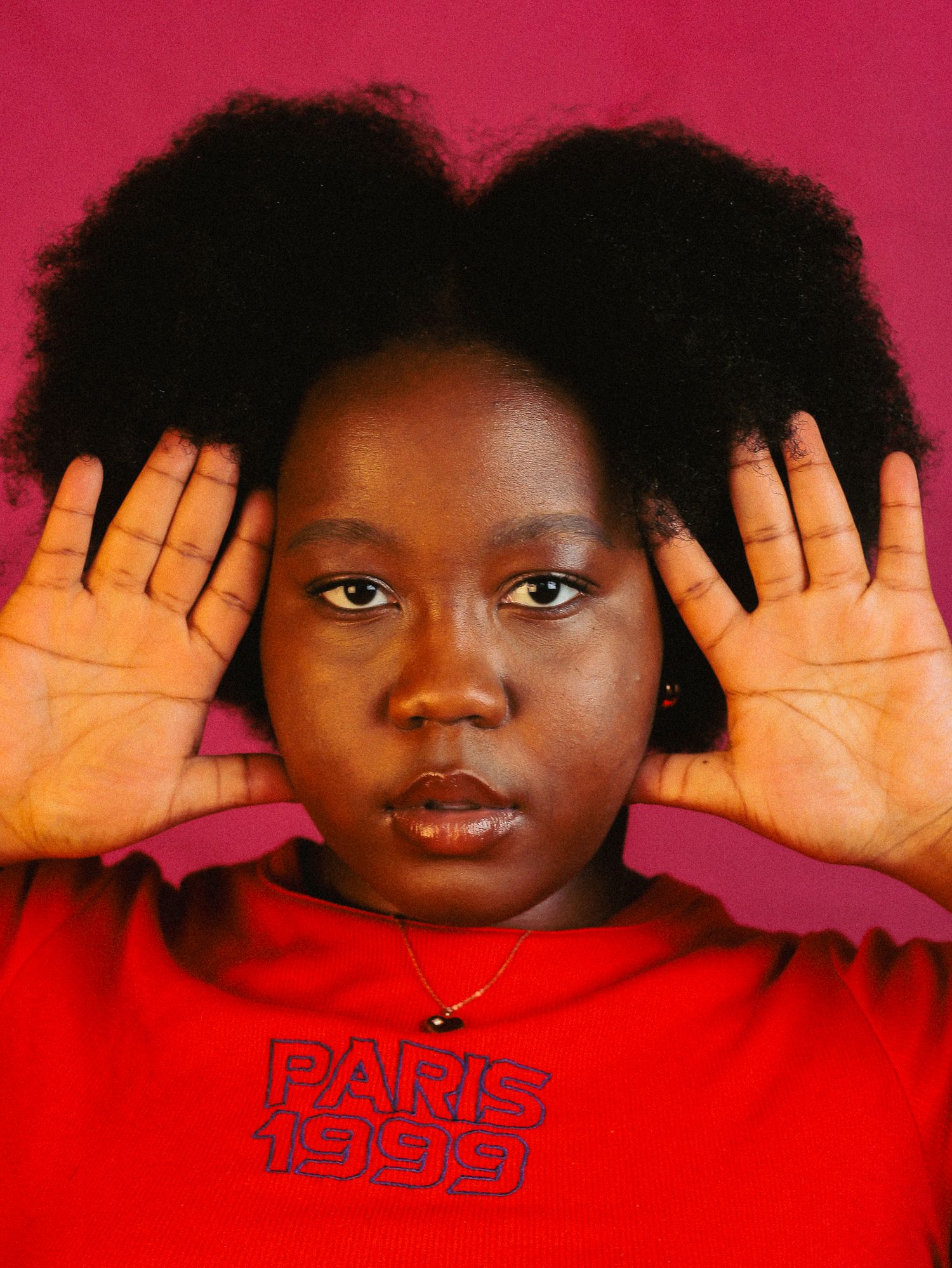 Black woman with natural afro hair holding hands up beside face, wearing red Paris 1999 shirt, pink background.