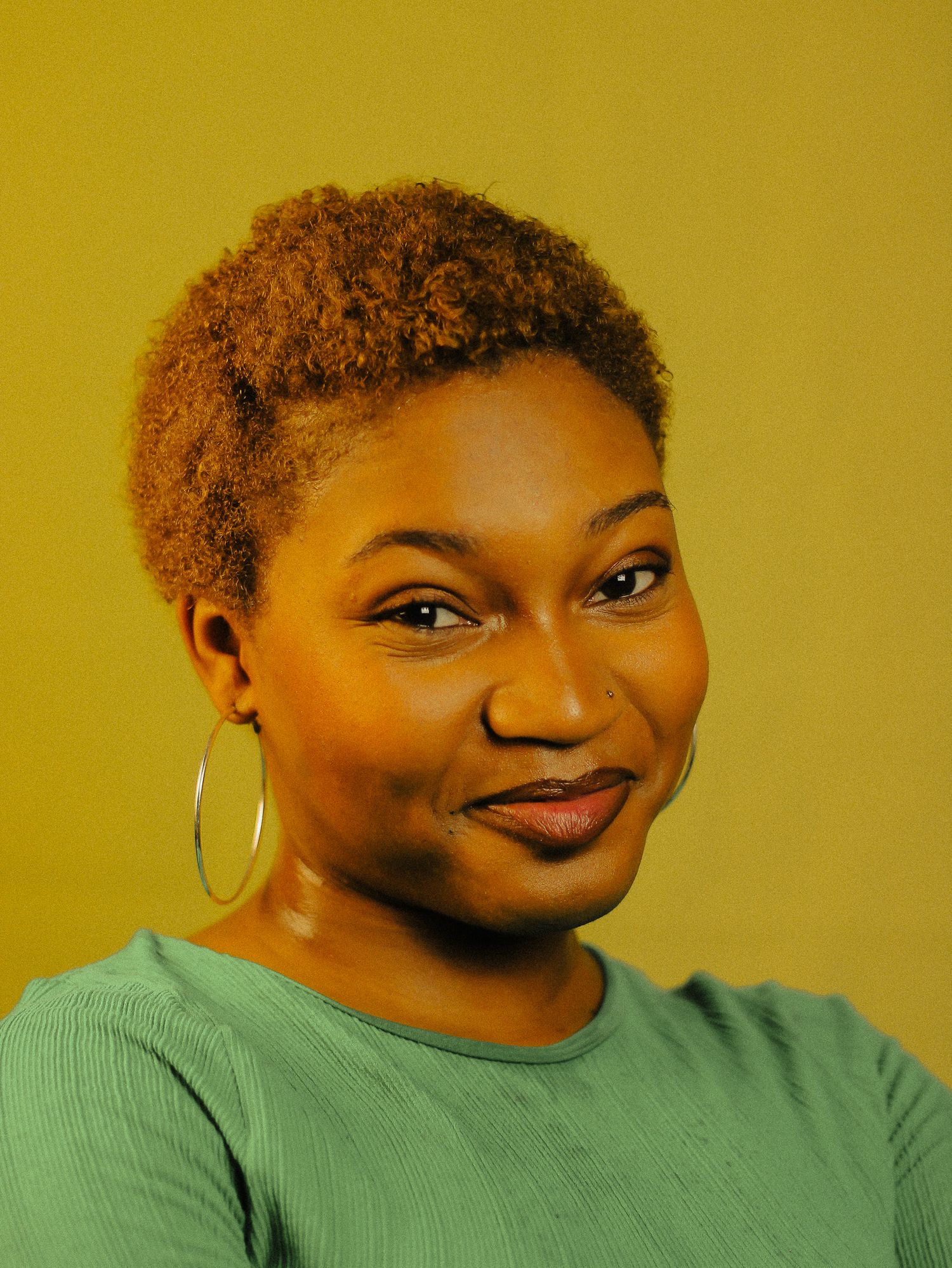 A young Black woman with short natural auburn hair smiles softly in a green top on yellow background.