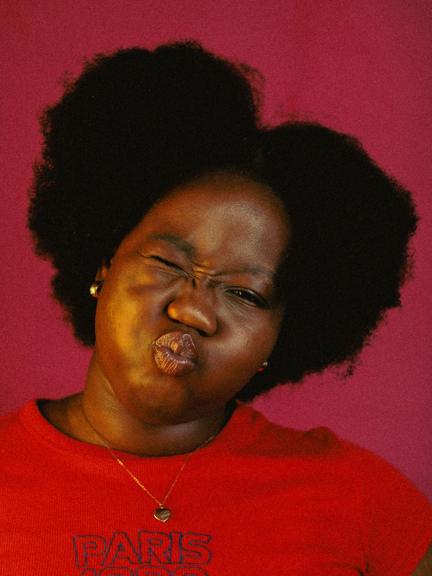 Young Black woman with afro hair making funny face in red Paris shirt on pink background.
