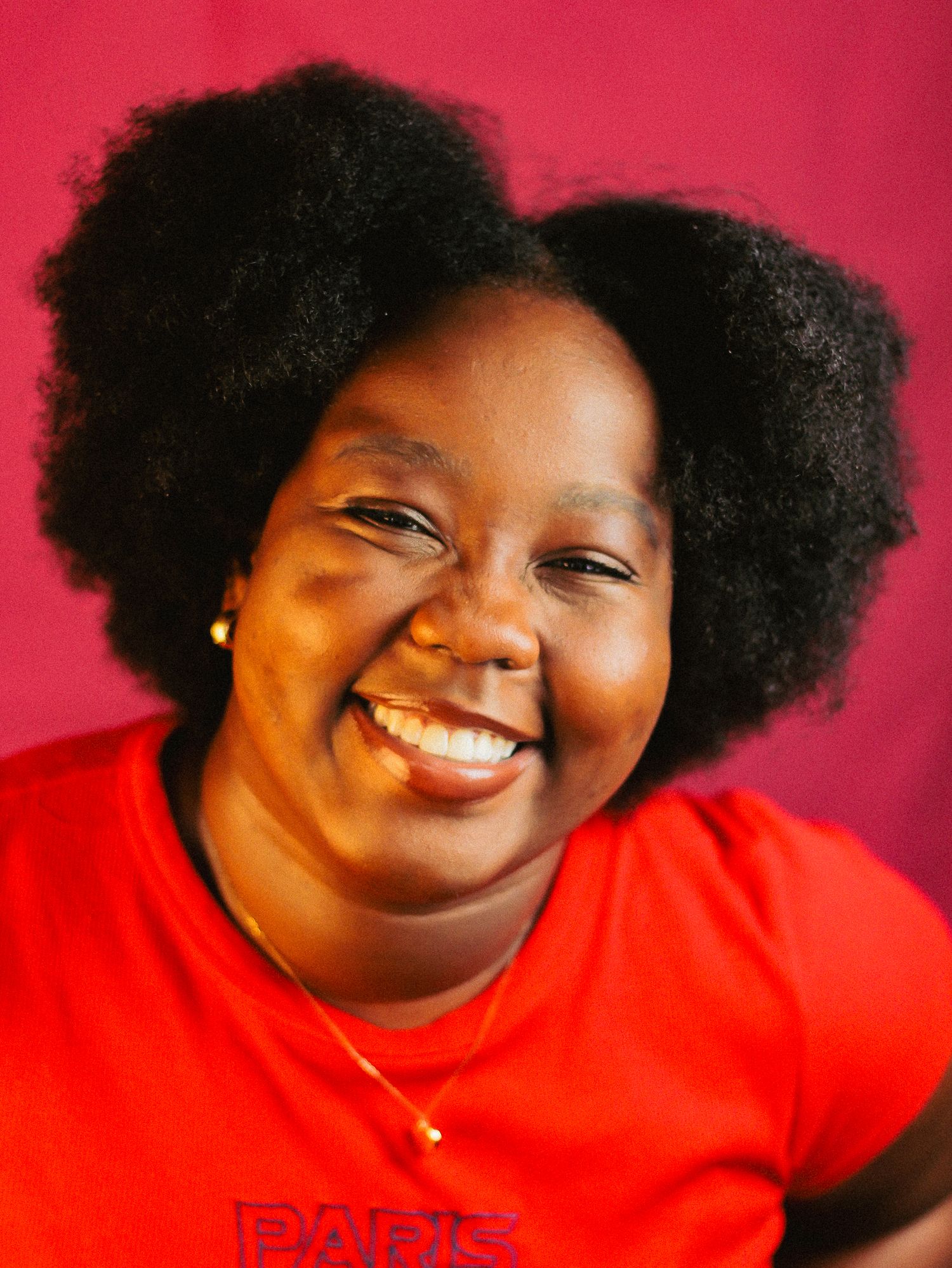 Cheerful Black woman with natural afro hair laughing in a red Paris t-shirt against a dark pink background.