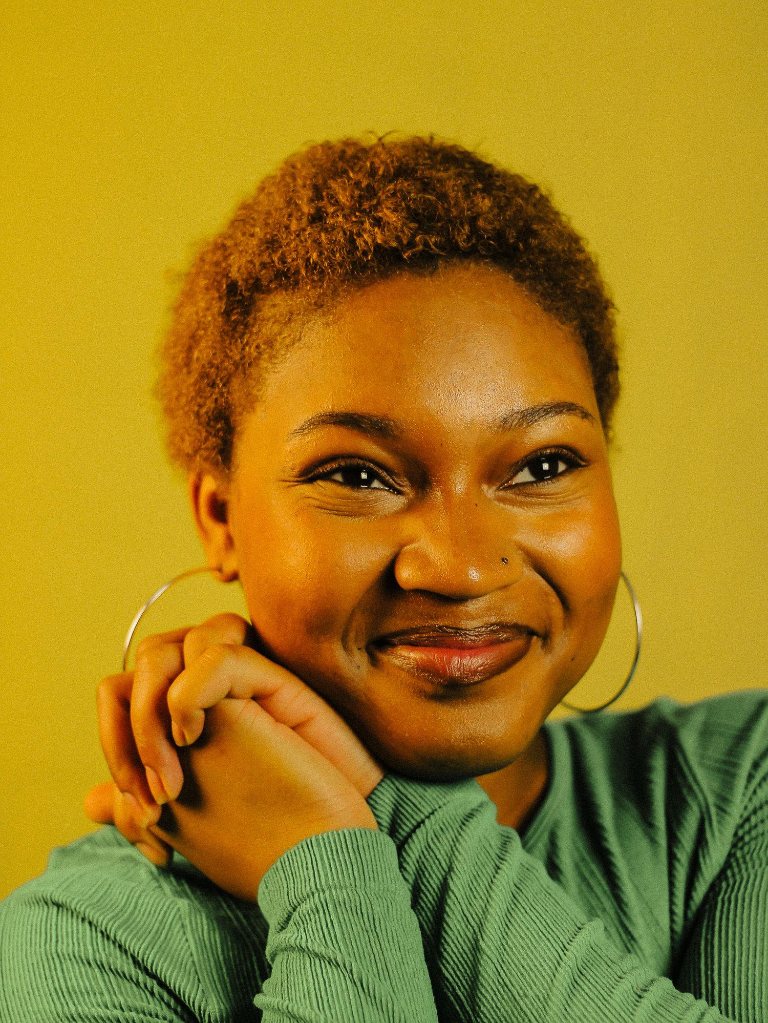Young woman with short curly hair smiling warmly with hands clasped under chin, wearing teal top and hoop earring.