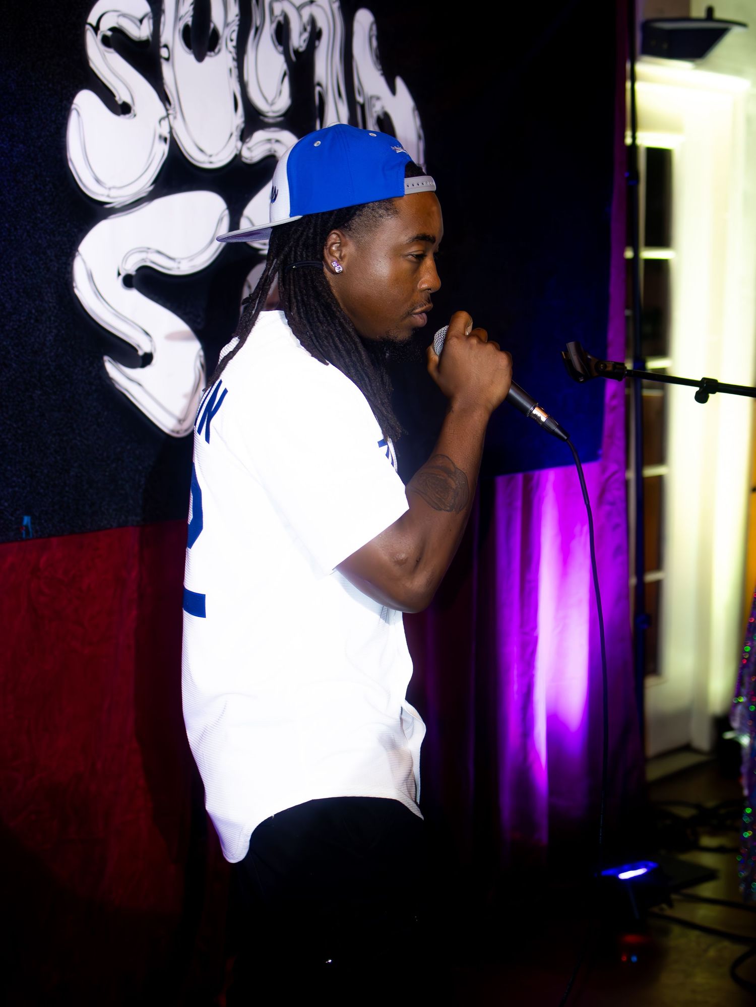 Performer in white shirt and blue cap singing on stage with artistic backdrop and mood lighting.