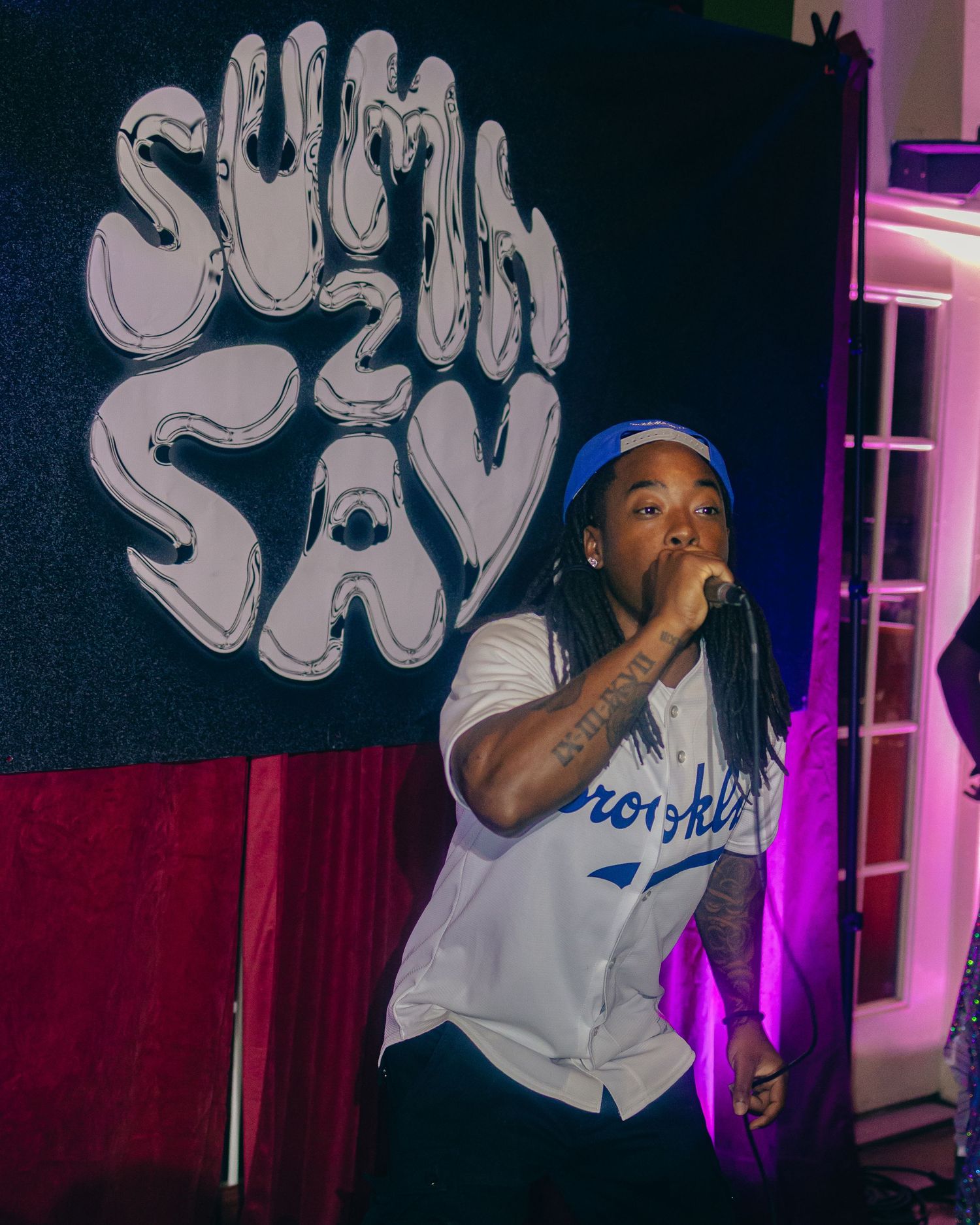 Performer in baseball jersey raps on stage with urban art backdrop and colored lights.