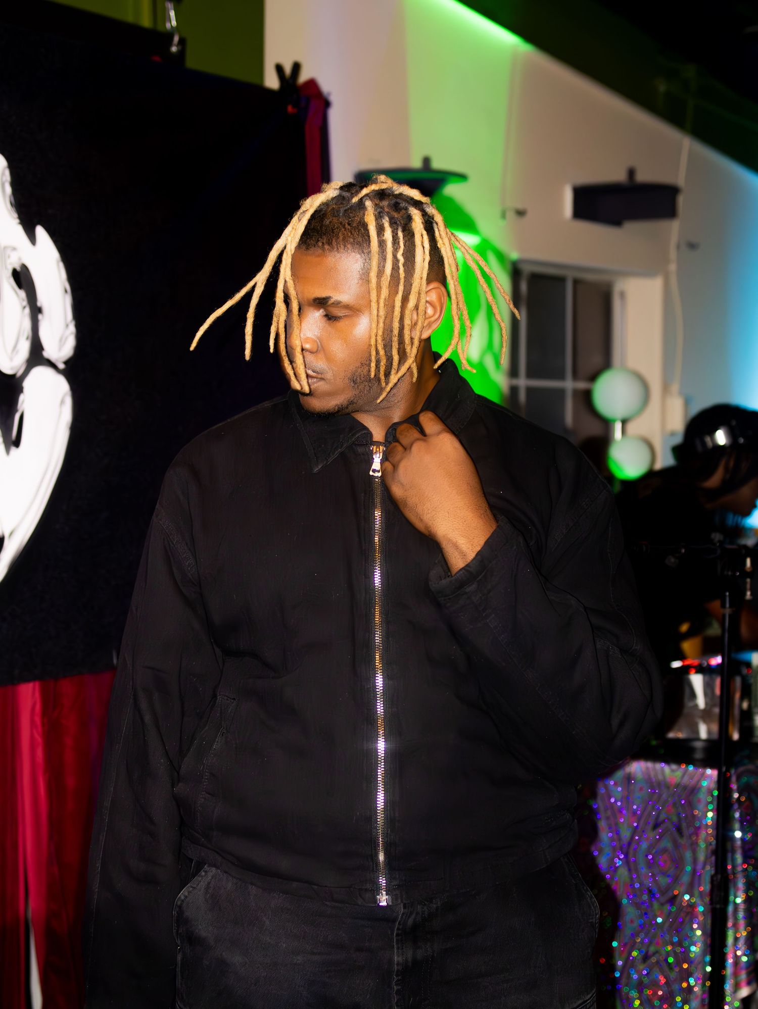 Performer in black jacket with blonde braids singing on stage with colorful lighting effects.