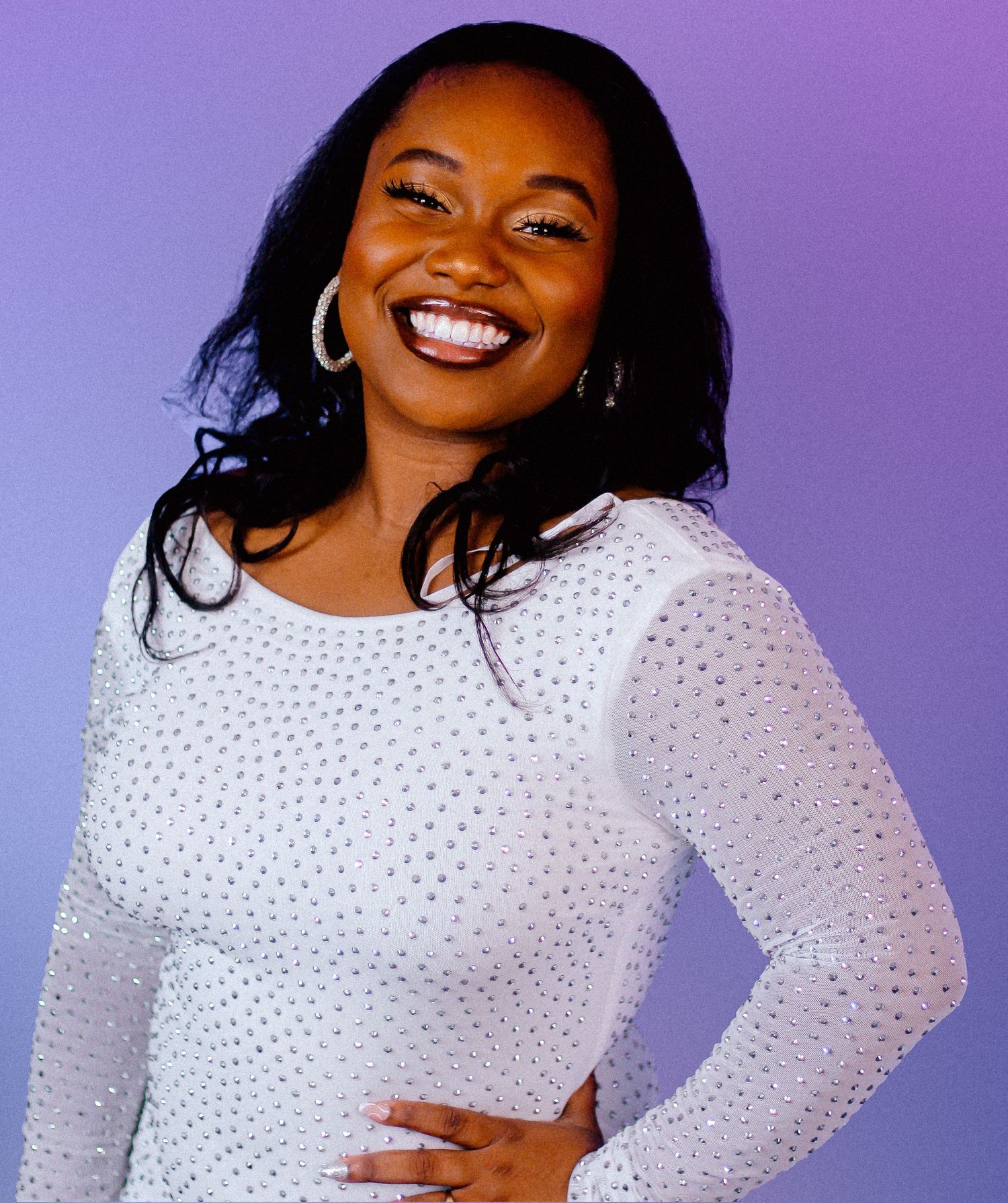 Portrait against purple backdrop showing sparkly white long sleeve top and joyful expression.