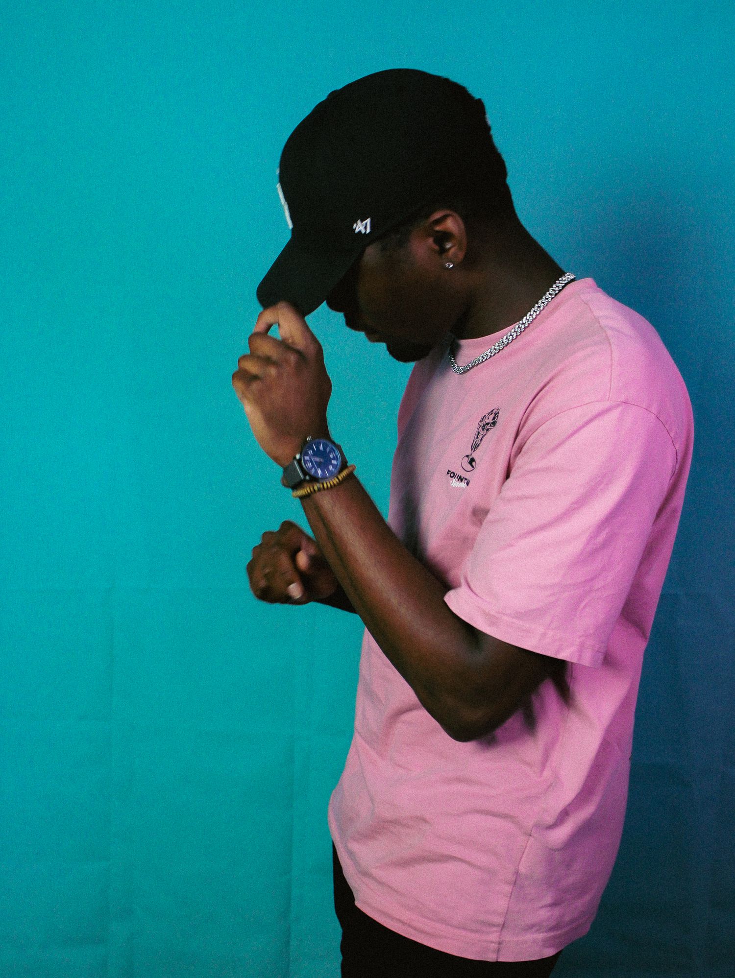 A young man in a pink t-shirt and black cap adjusts his hat against a teal wall.