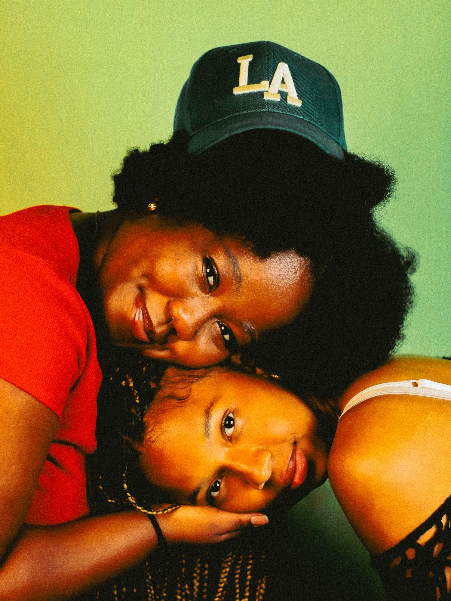 Two Black women in affectionate pose, one with natural afro leaning over other wearing navy LA cap smiling.