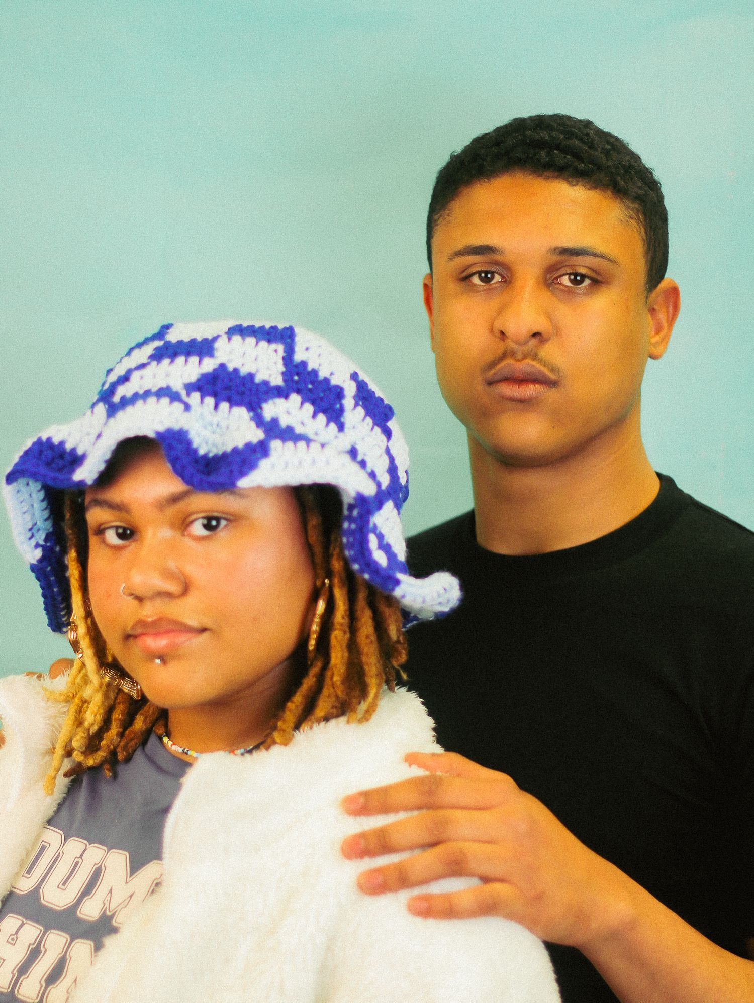 Young woman in blue crochet bucket hat and fluffy jacket poses with young man in black tee on teal background.