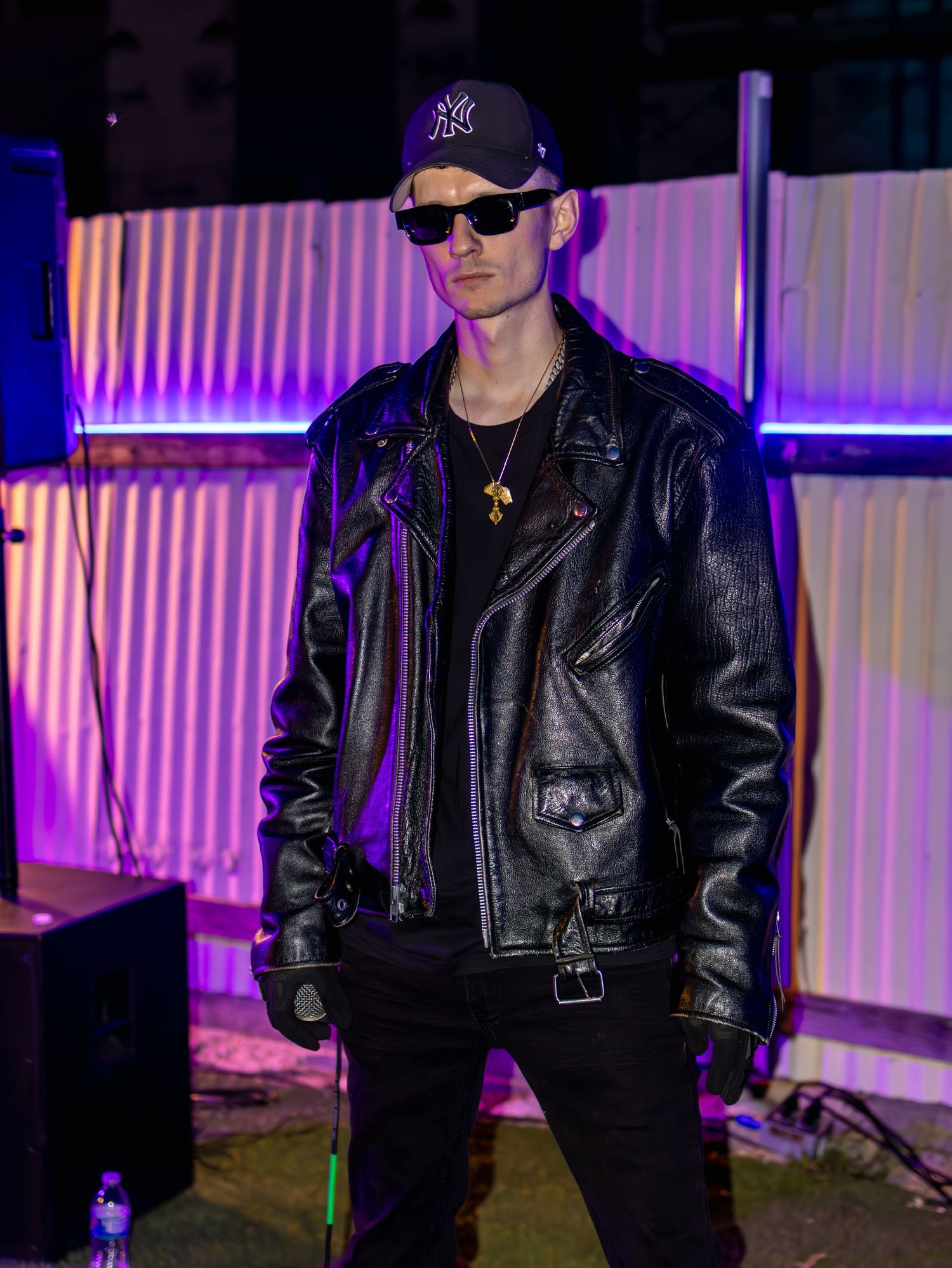 Person in black leather jacket and baseball cap performing under dramatic purple lighting.