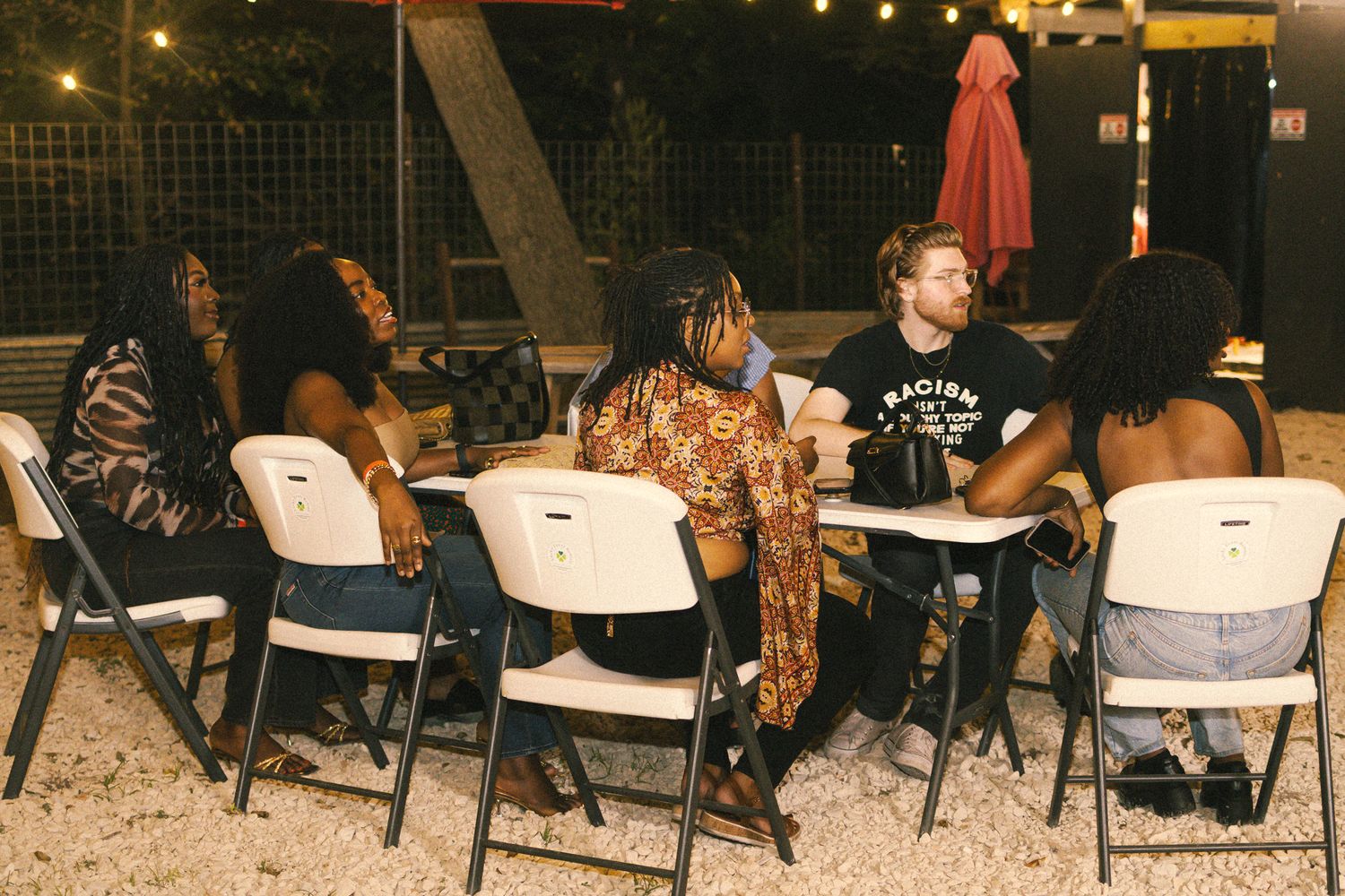 Row of white tables and chairs set up outdoors under string lights at night.