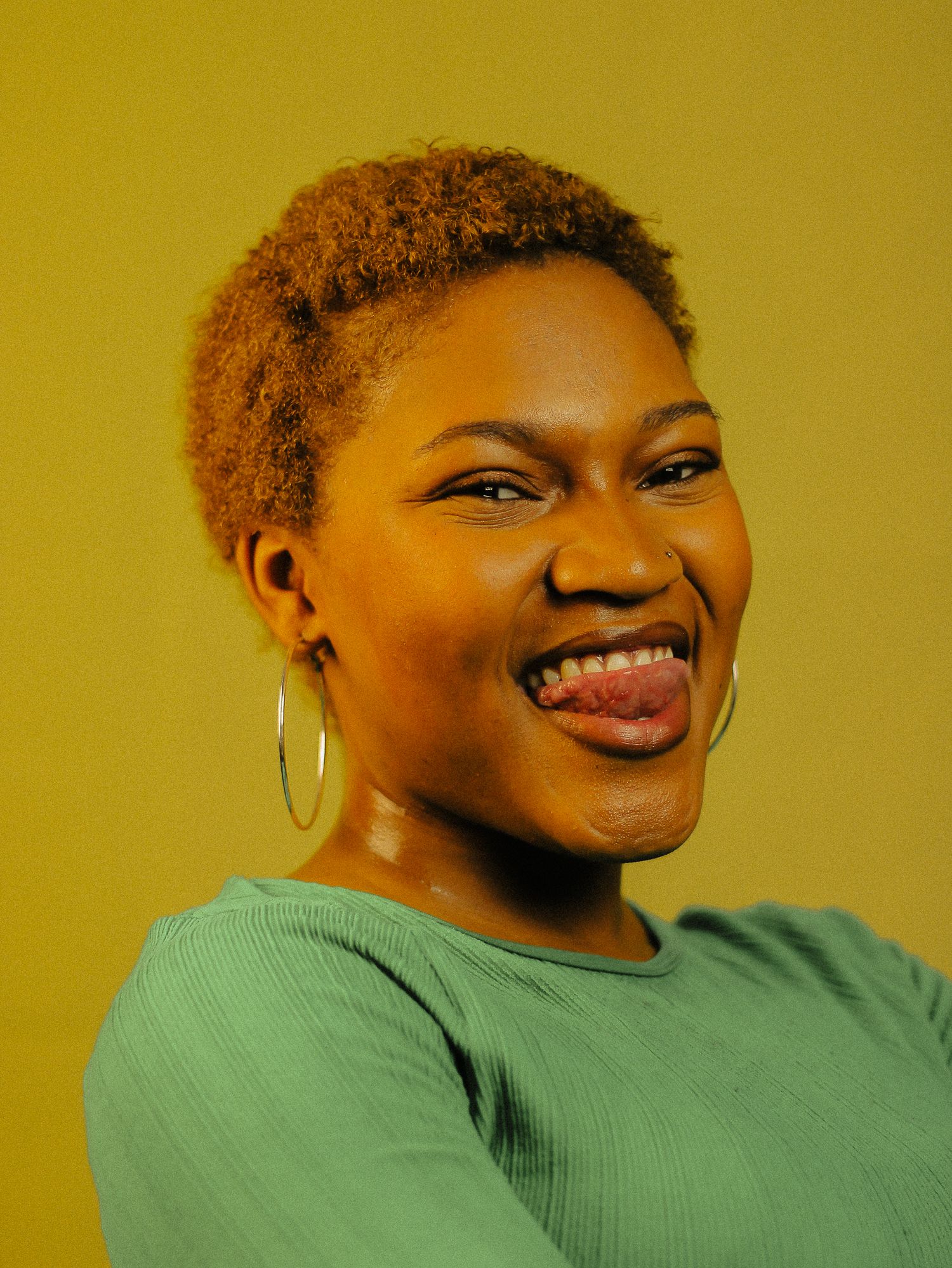 A playful young Black woman in a green top sticks out her tongue against a warm yellow background.