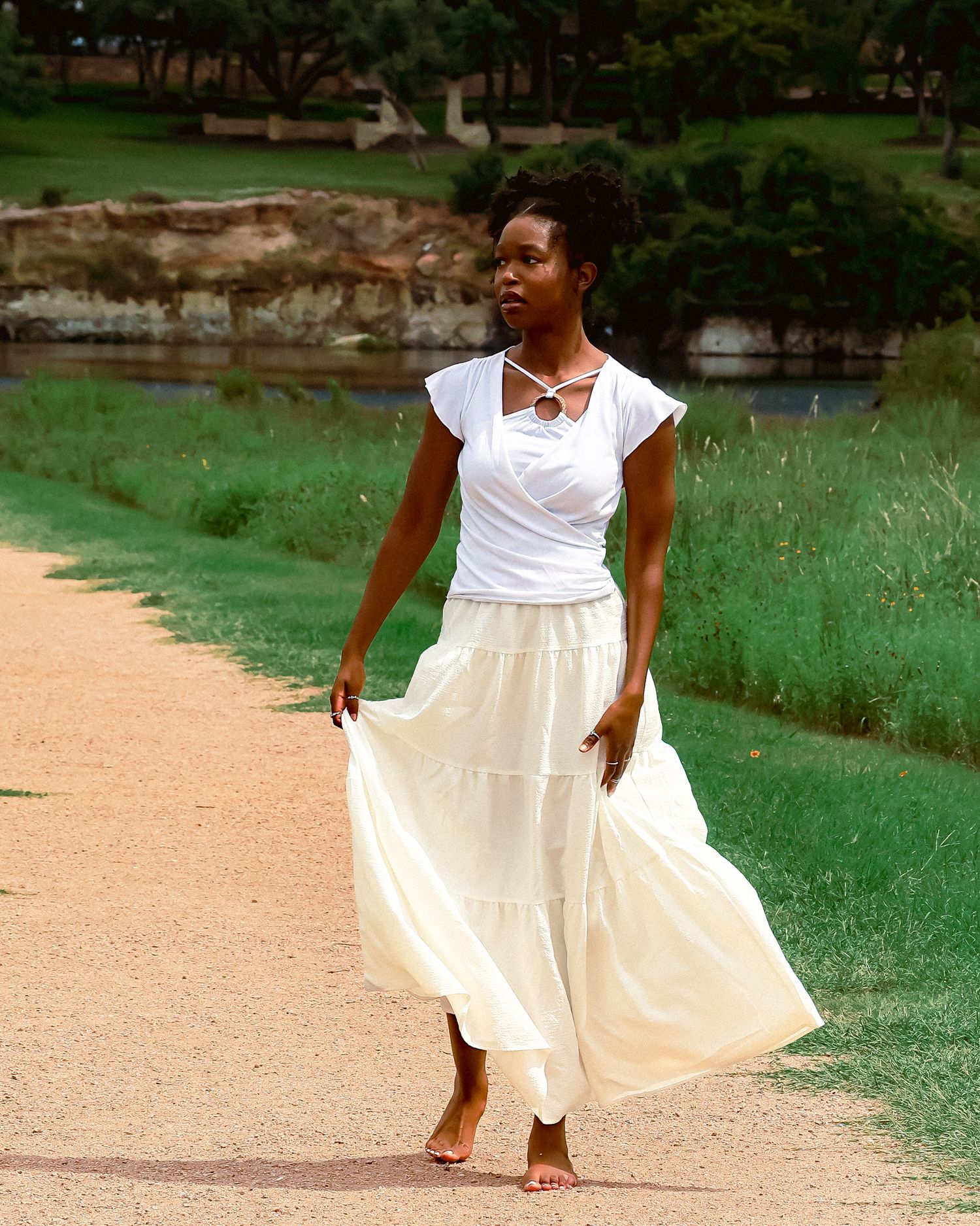 Woman in white summer dress walking barefoot along path near water.