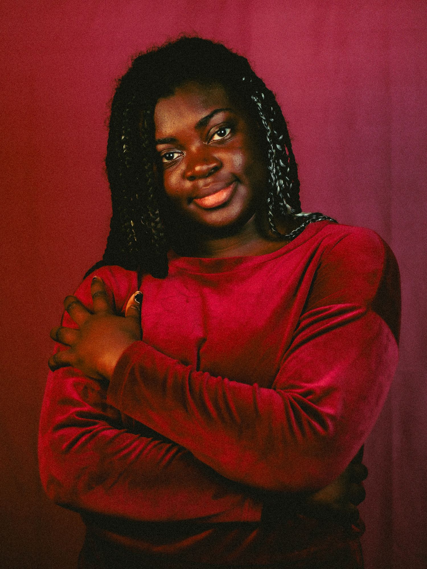 Young woman in a red velvet top poses with arms crossed, smiling confidently against a dark red background.