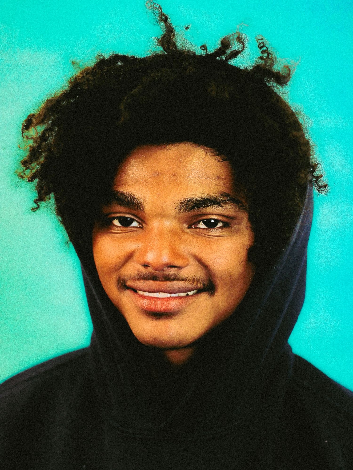 Young man with curly hair smiling warmly wearing dark hoodie against turquoise background.